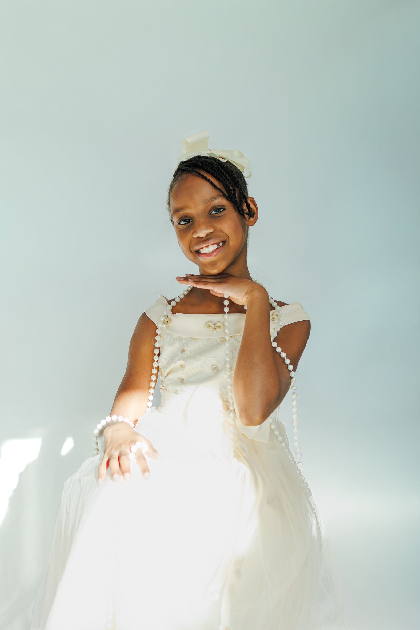 Happy young Black girl in white dress and pearl necklace posing with hands outstretched.