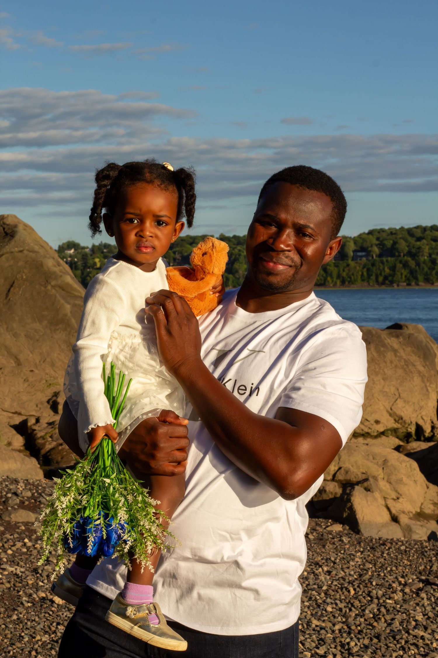 Smiling Black father holding young daughter with bouquet and stuffed animal by rocky waterfront.