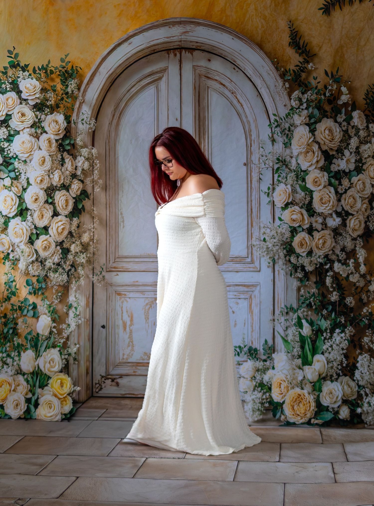 Woman in elegant white off-shoulder gown poses by ornate door with white roses.