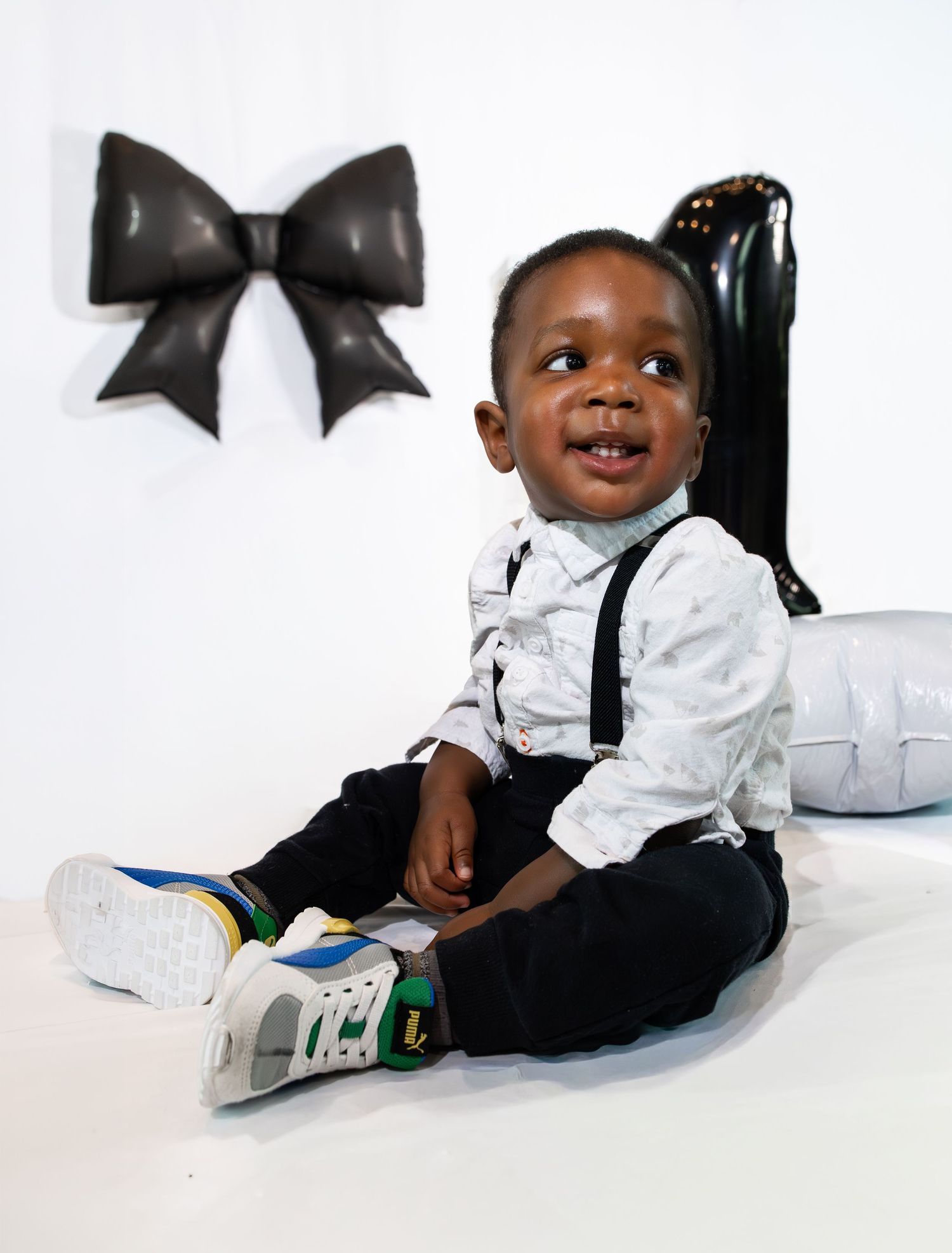 Smiling toddler in suspenders, white shirt, black pants, and colorful sneakers sits on floor near decorative black bow.