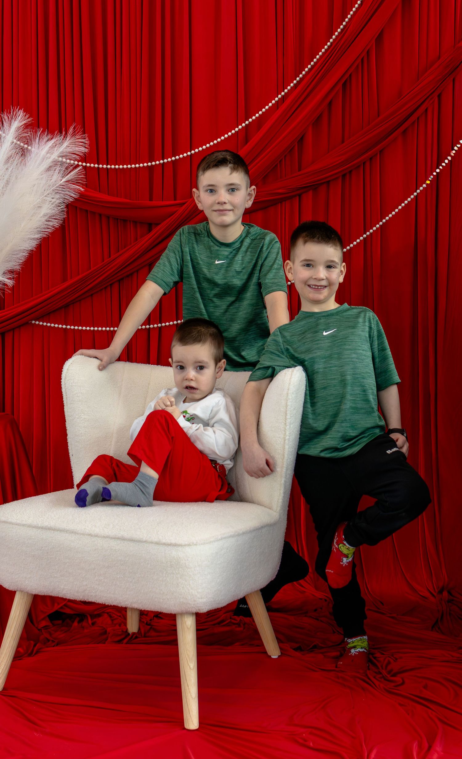 Three boys pose for a holiday photo in front of red curtains, two older boys in green Nike shirts with a baby in a chair.