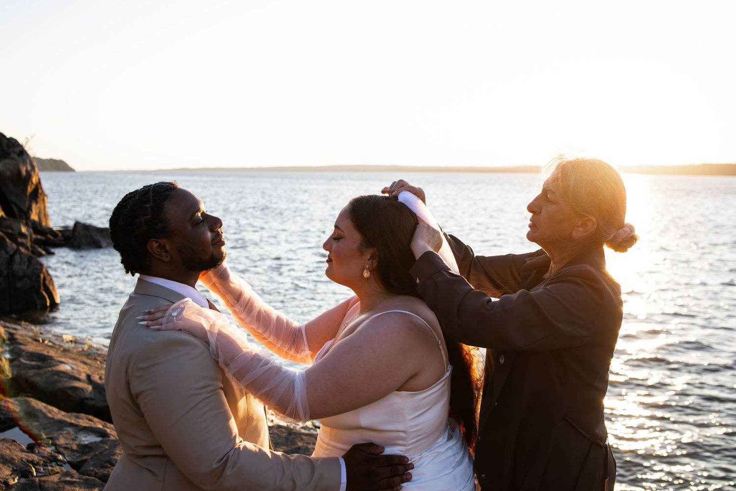 Couple exchanges vows at sunset on rocky shoreline with officiant adjusting brides hair during outdoor wedding ceremony.