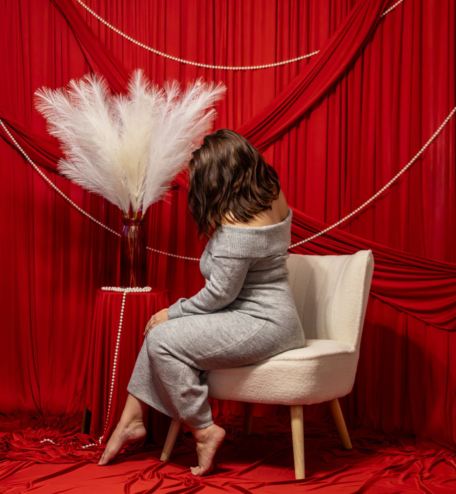 Woman in grey outfit sitting with her back to camera beside white pampas grass on red backdrop.