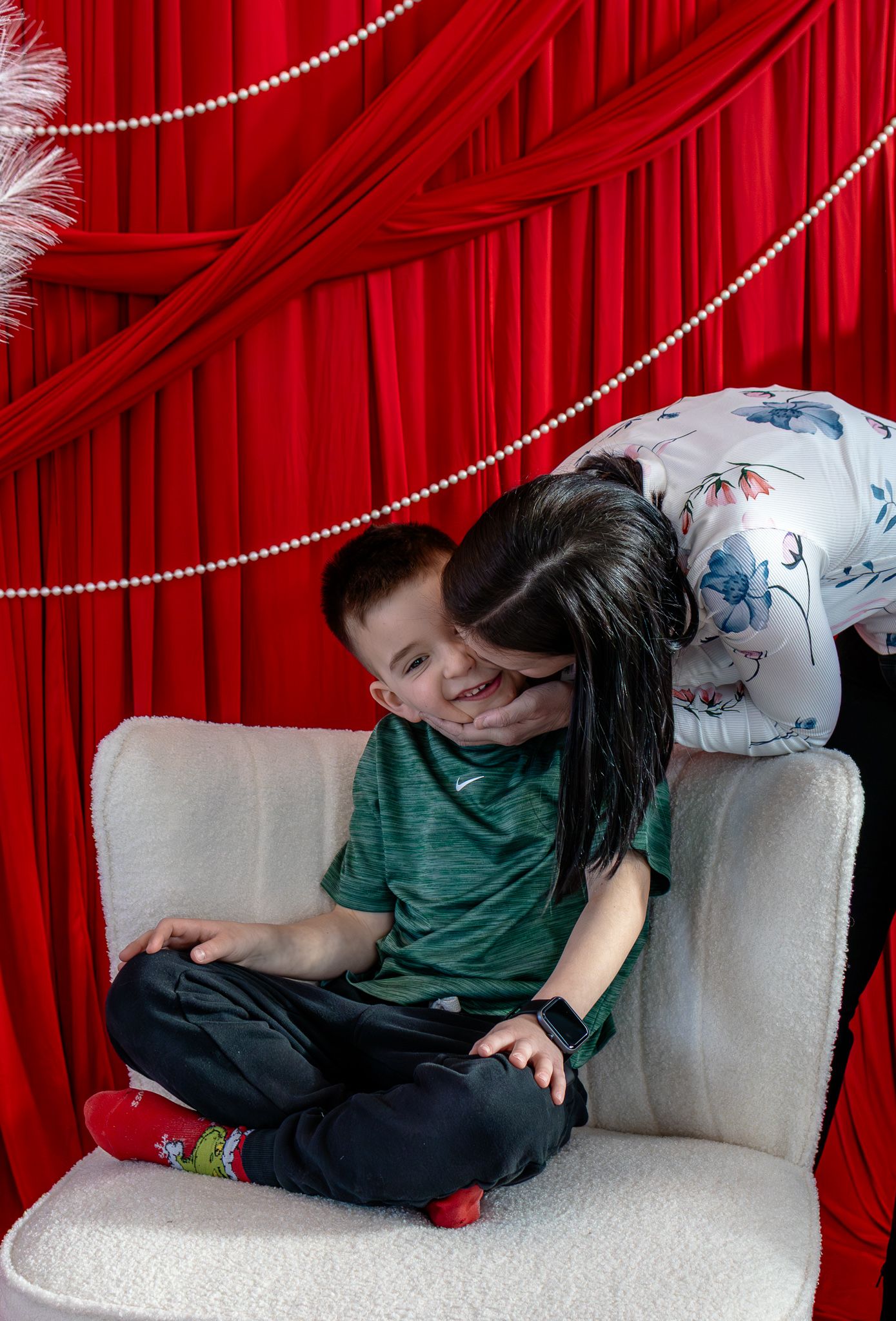 Woman hugging a smiling boy sitting cross-legged on a white chair against red curtain backdrop.