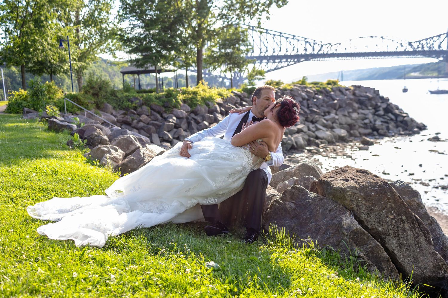 Bride and groom share a romantic kiss on rocky shoreline with bridge and water in background on sunny wedding day.