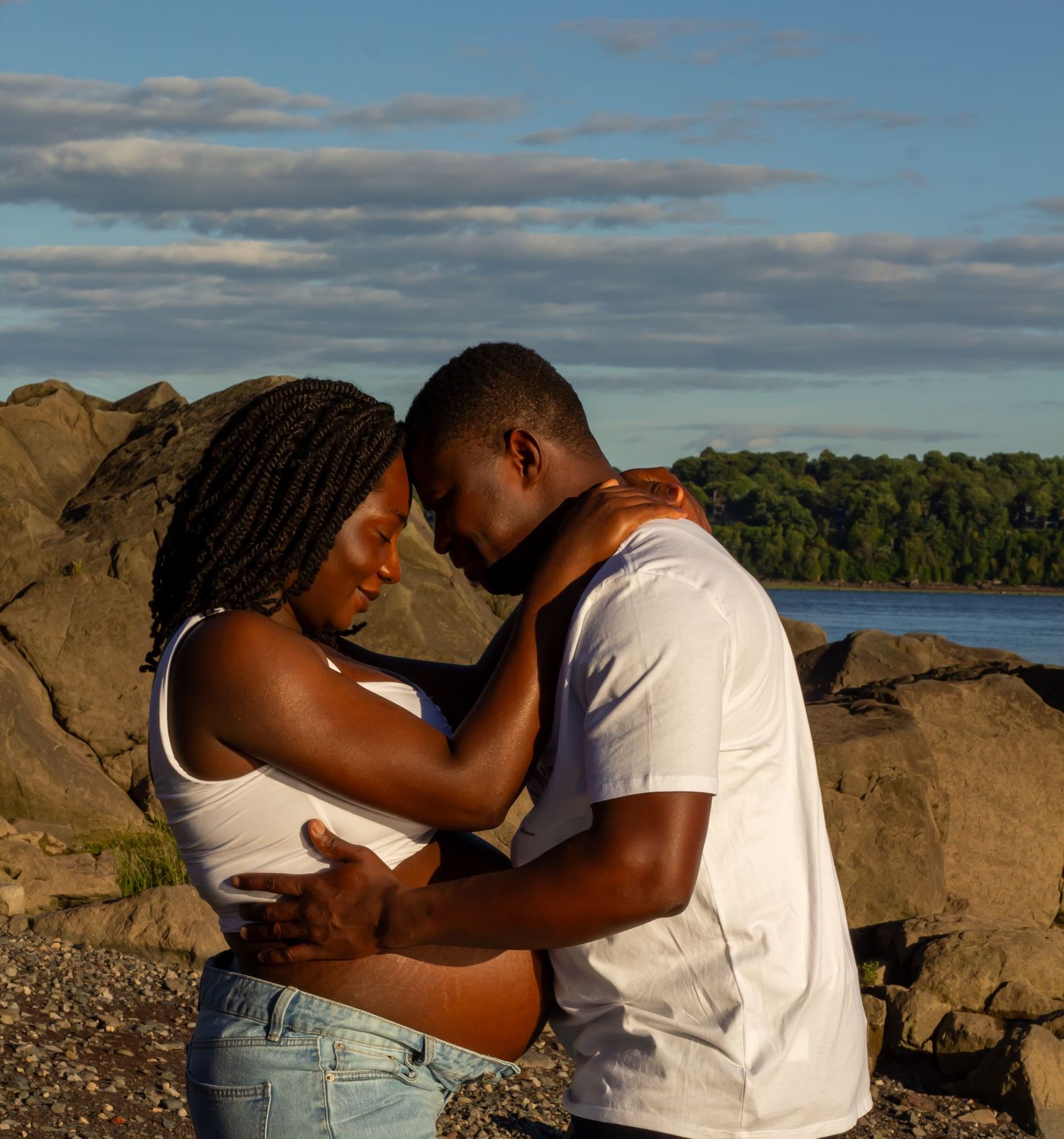 Black couple sharing intimate forehead-to-forehead embrace on rocky beach at golden hour.