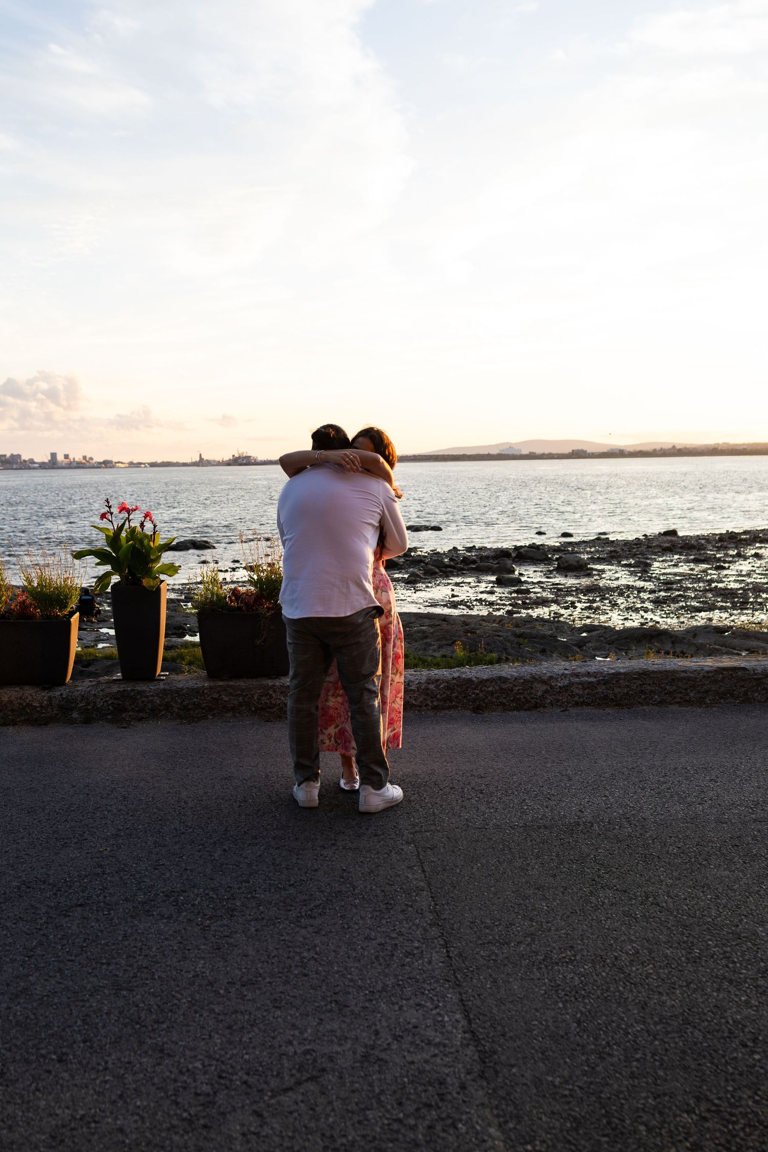 Couple embracing by the waterfront at sunset, with flowers and a city skyline in the background.