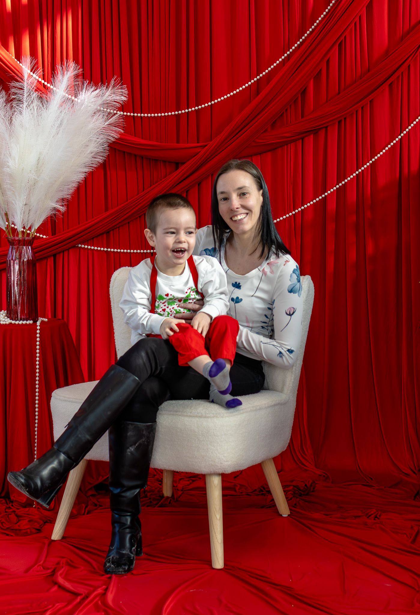 Smiling mother and toddler son sitting together on a white chair with festive red curtain backdrop.