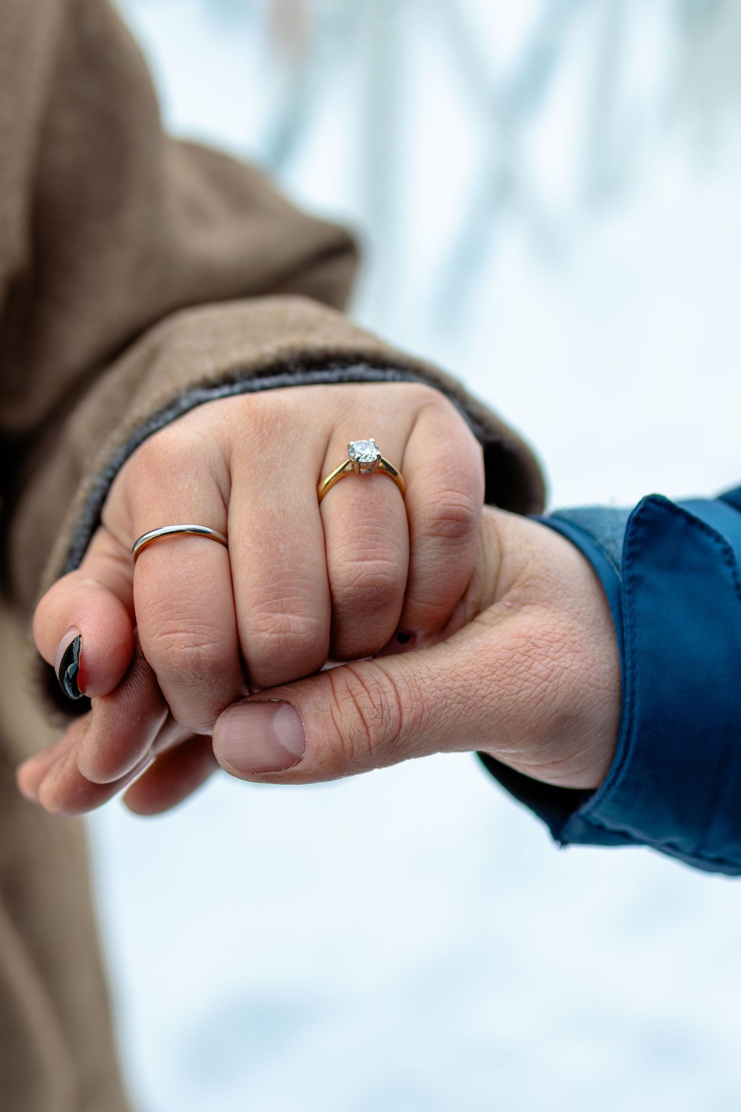 Close-up of two hands holding, woman wearing gold diamond engagement ring and silver band, snowy winter background.