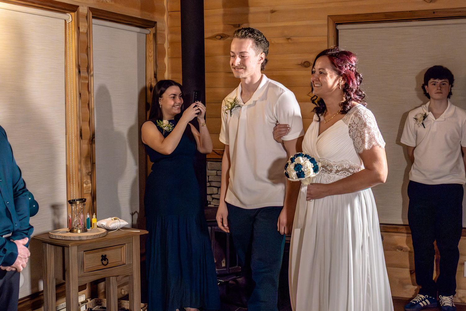 Bride and groom stand together during wedding ceremony while guest photographs the moment in a rustic wooden venue.