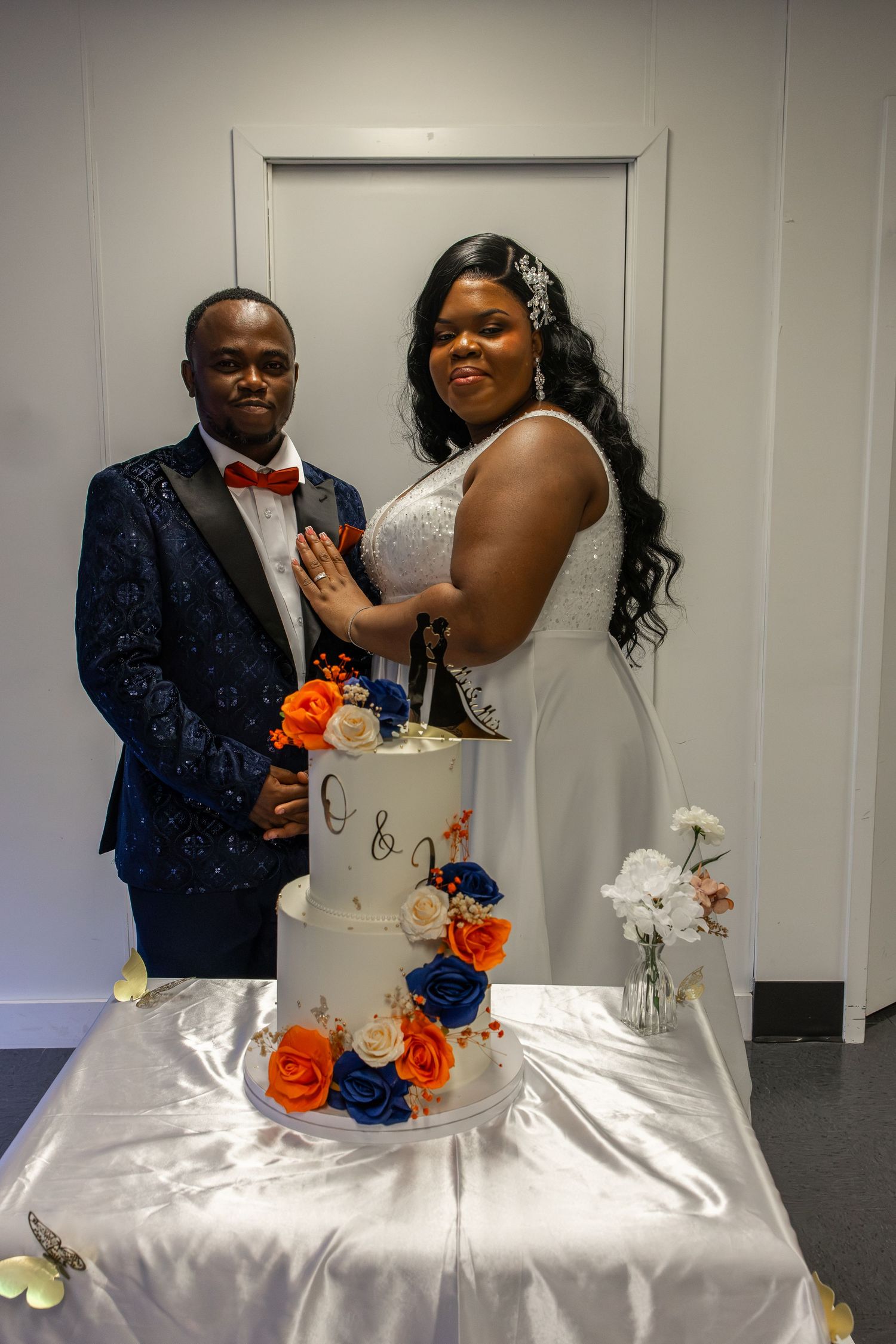 Newlywed couple cutting a two-tier white wedding cake decorated with orange and blue roses at their reception.