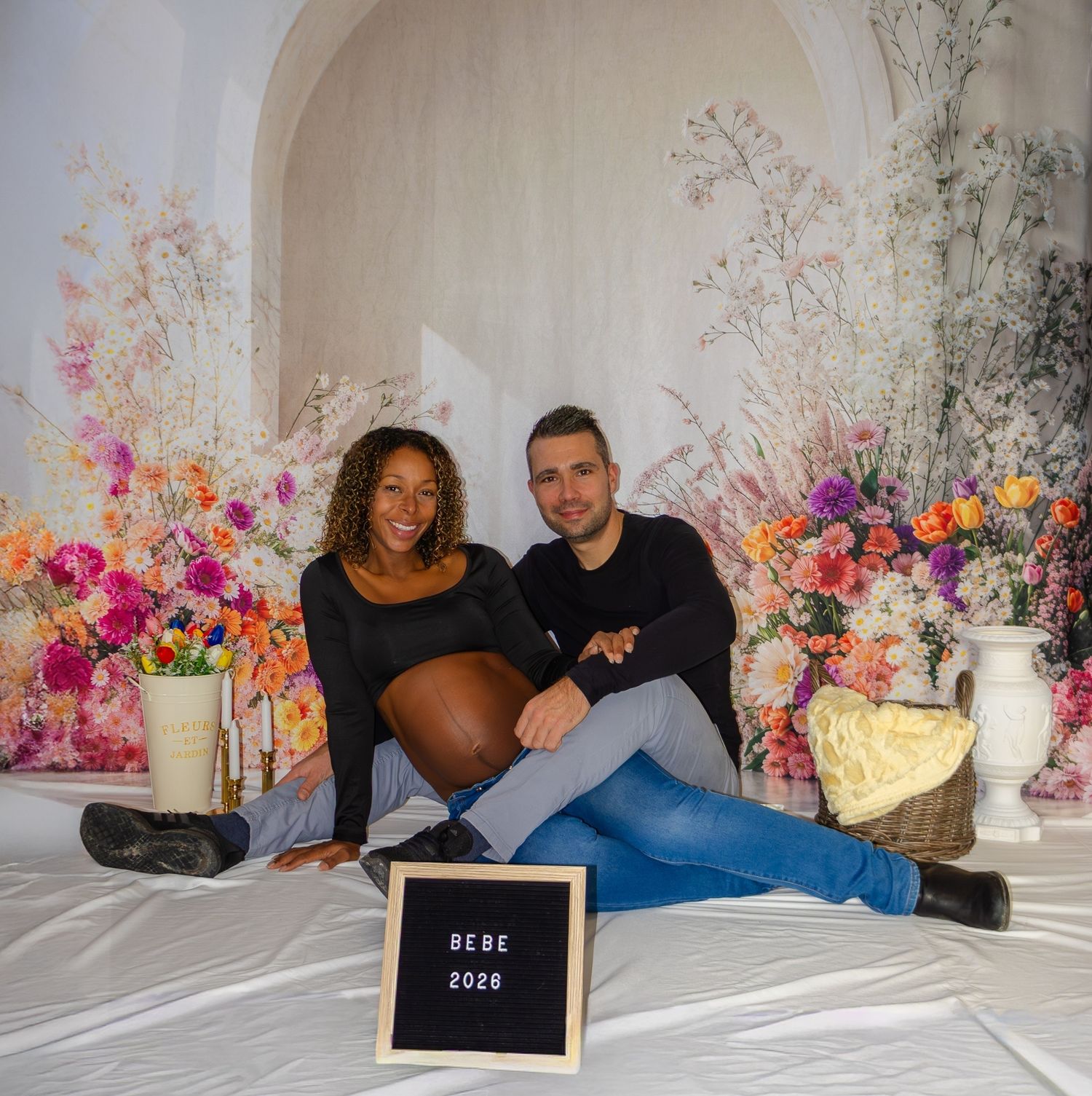 Interracial couple sitting on floor with baby bump and BEBE 2026 sign amid colorful floral backdrop.