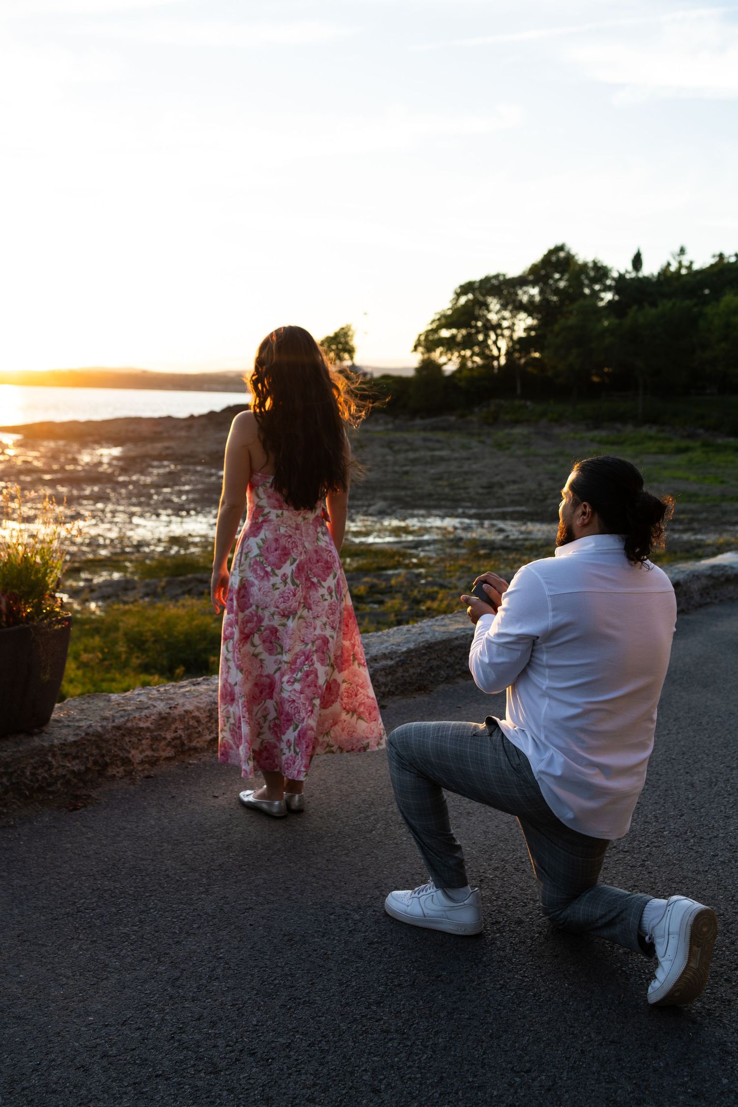 Man kneeling on one knee proposing to woman in floral dress at sunset by the water.