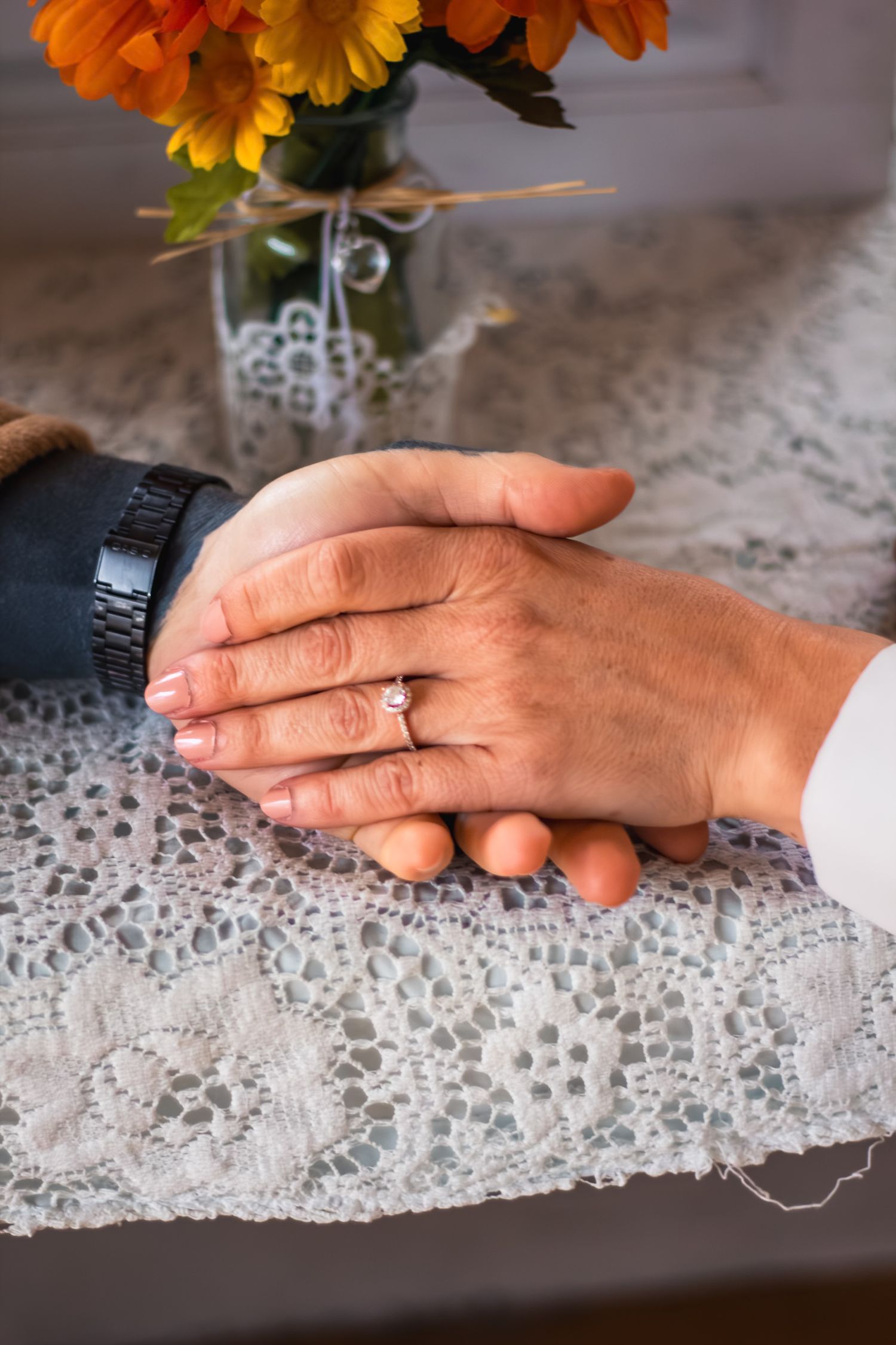 Close-up of couple holding hands on lace tablecloth, woman wearing diamond engagement ring, autumn flowers behind.