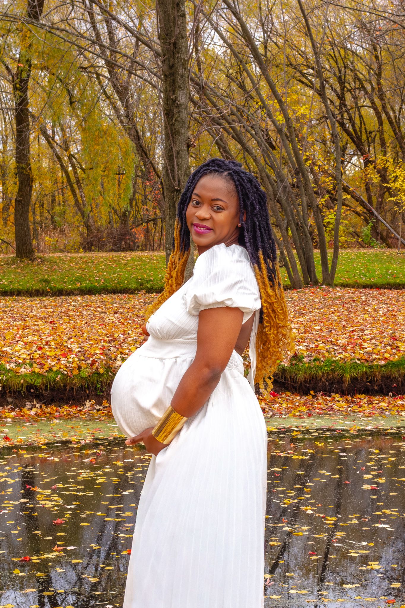 Pregnant African American woman in a white dress posing by a pond in an autumn park setting.