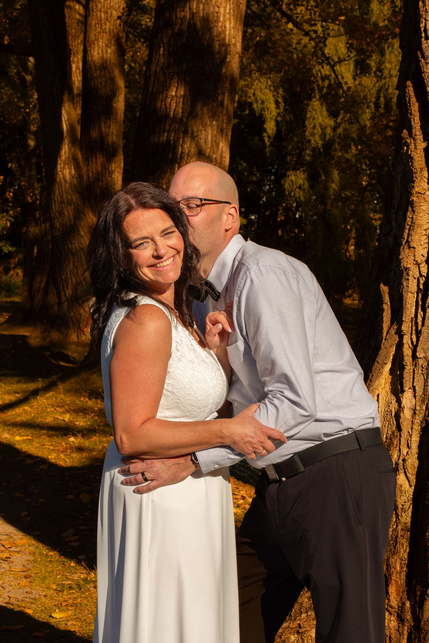 Couple sharing a romantic kiss in golden forest light, bride in white dress smiling joyfully.