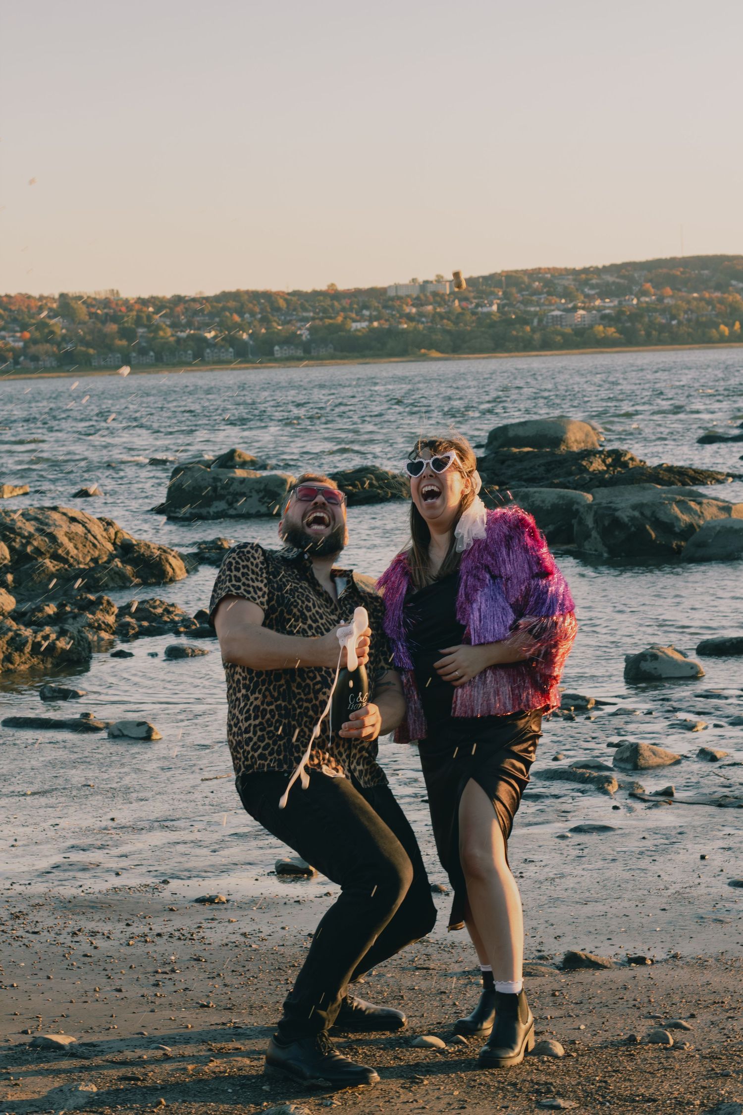 Two friends in funky outfits laugh and dance on a rocky beach at sunset with a city skyline in the background.