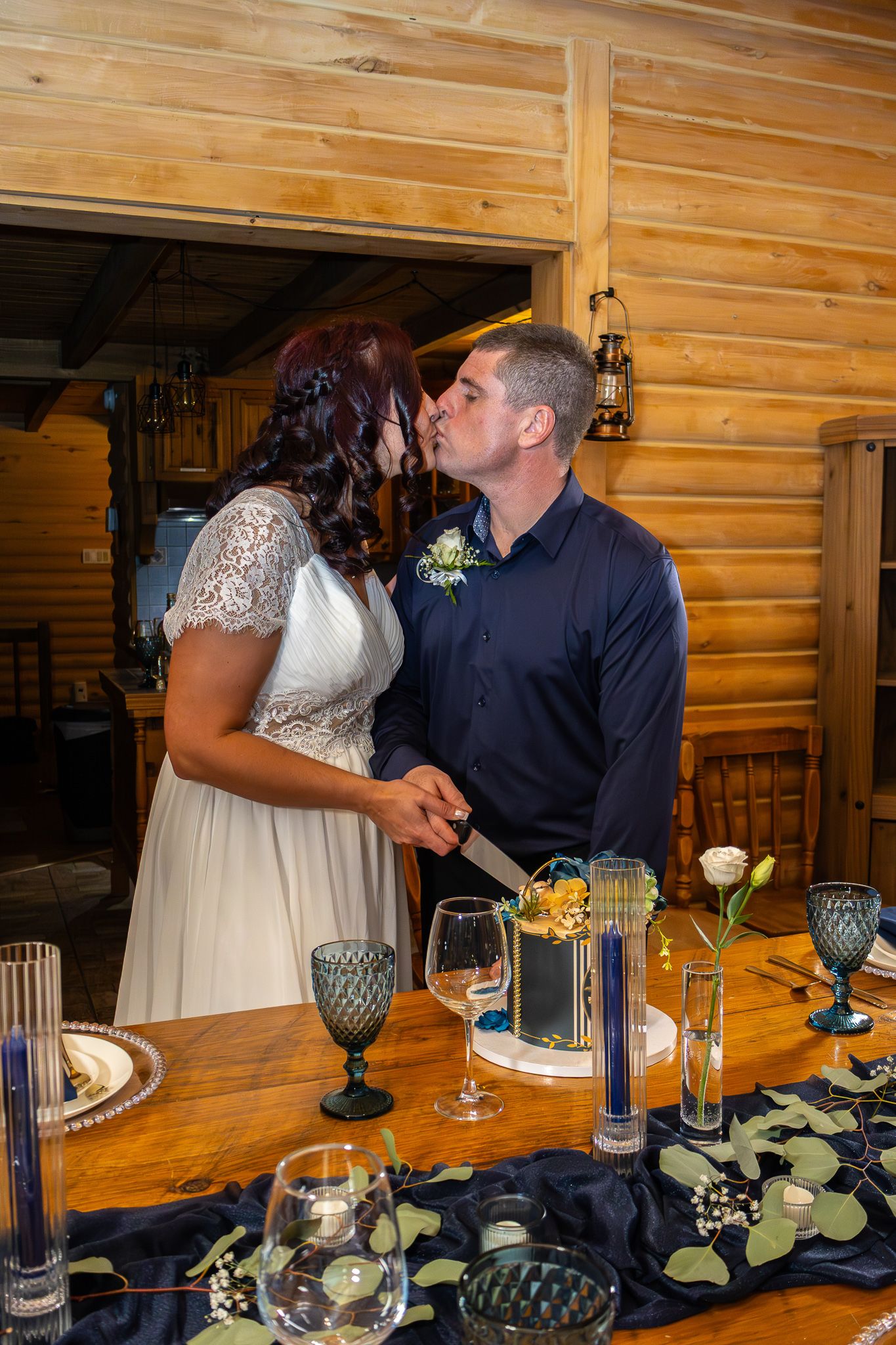 Bride and groom kiss while cutting their elegant navy and gold wedding cake at a rustic log cabin venue.