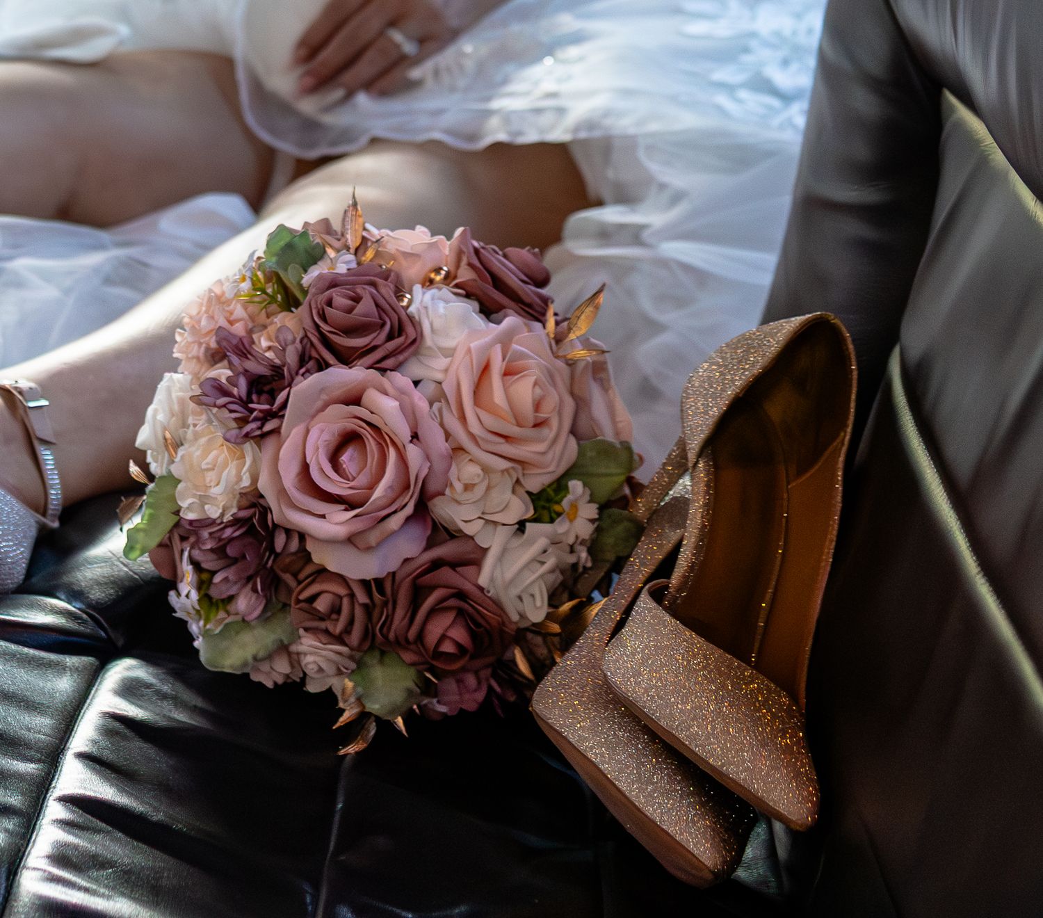 A bridal bouquet of mauve and pink roses beside gold glitter heels on a leather seat with a bride in white dress.