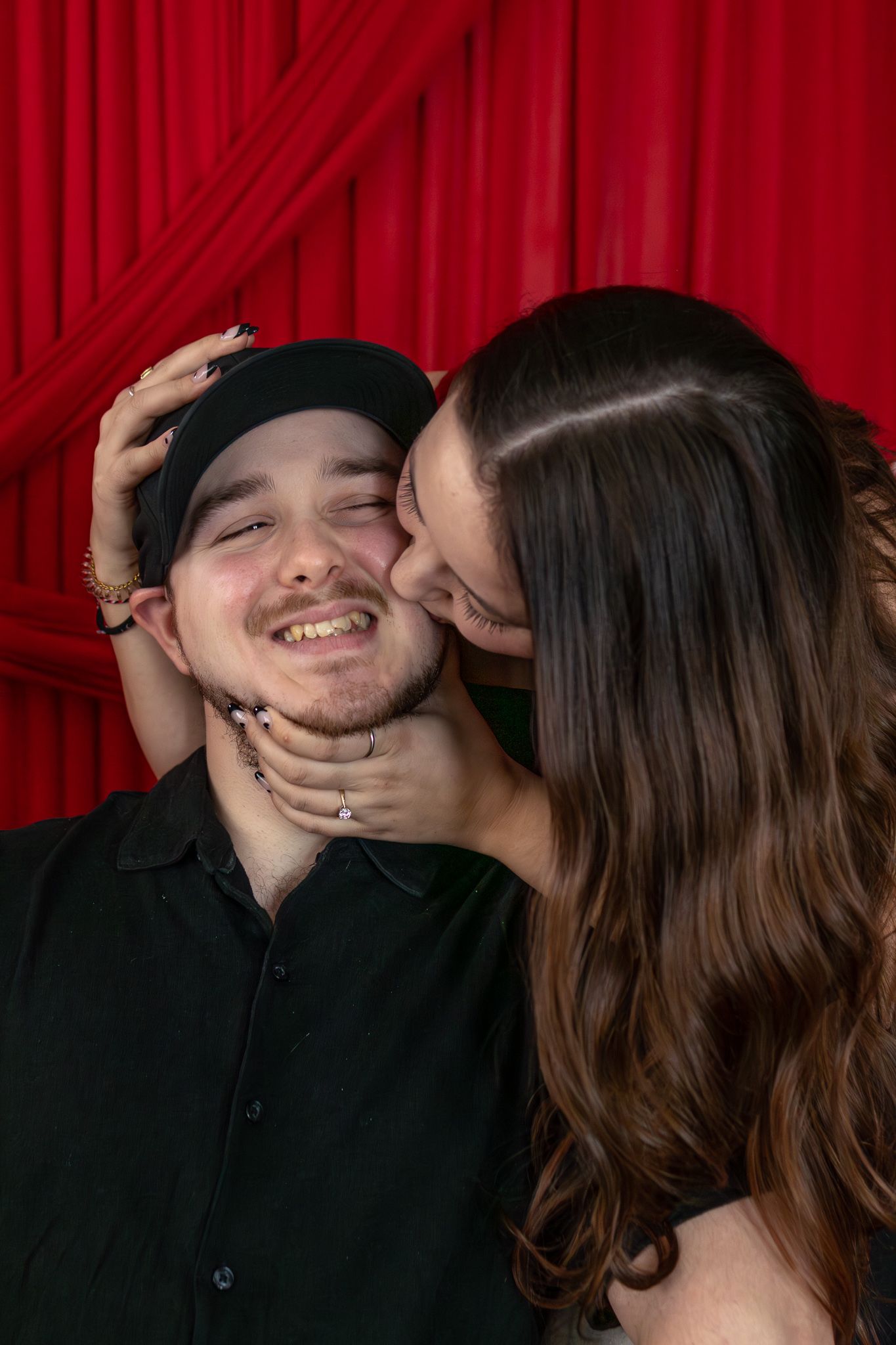Smiling man in black cap enjoys kiss on cheek from woman against red curtain background.