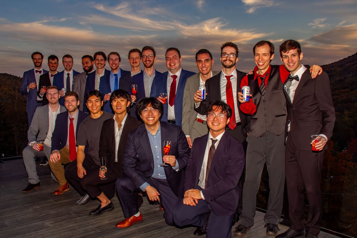 Large group of male wedding guests pose together smiling at an outdoor venue during a colorful sunset.