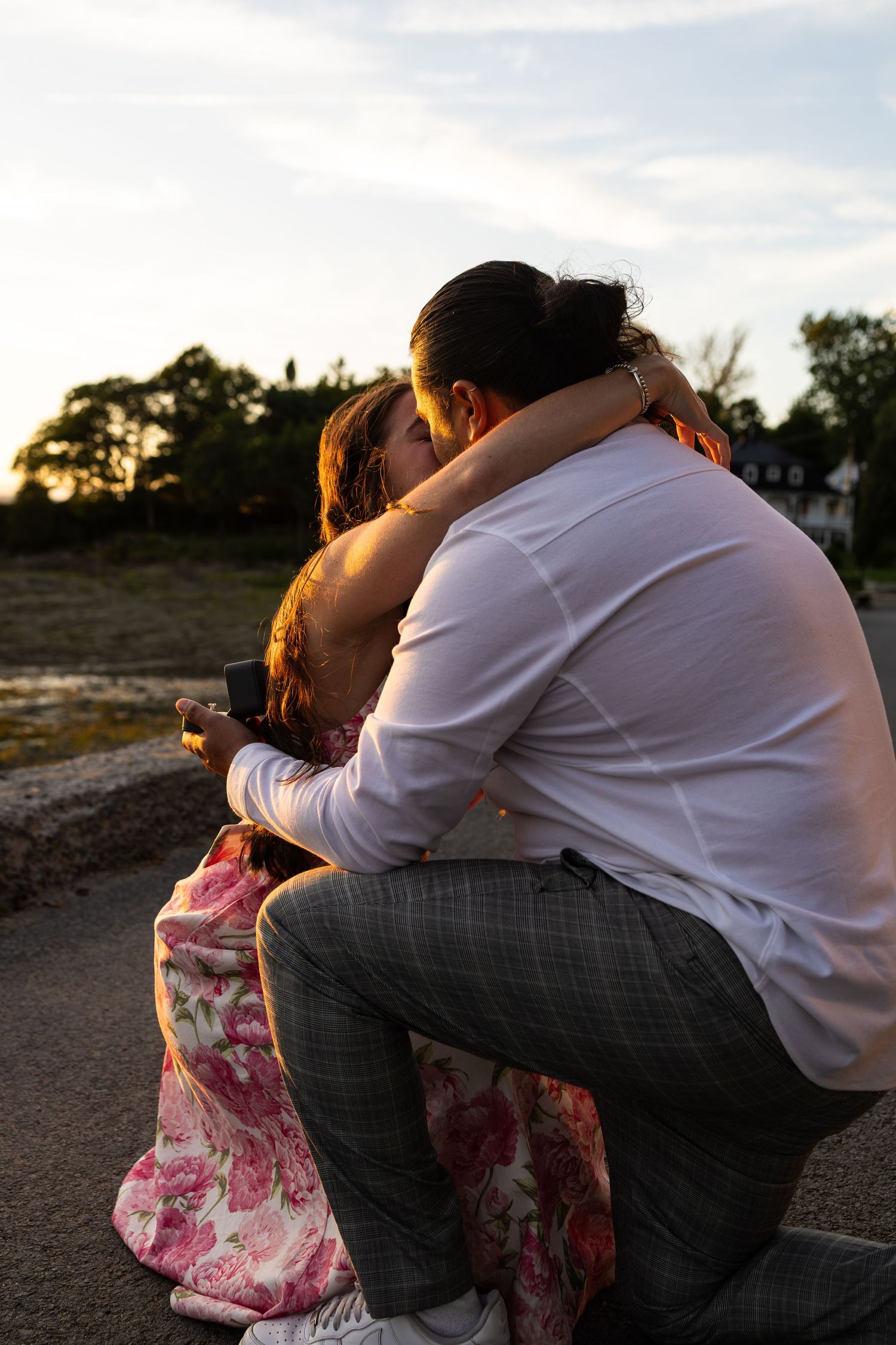 Couple sharing a romantic embrace at sunset, sitting outdoors with warm golden light and trees in the background.