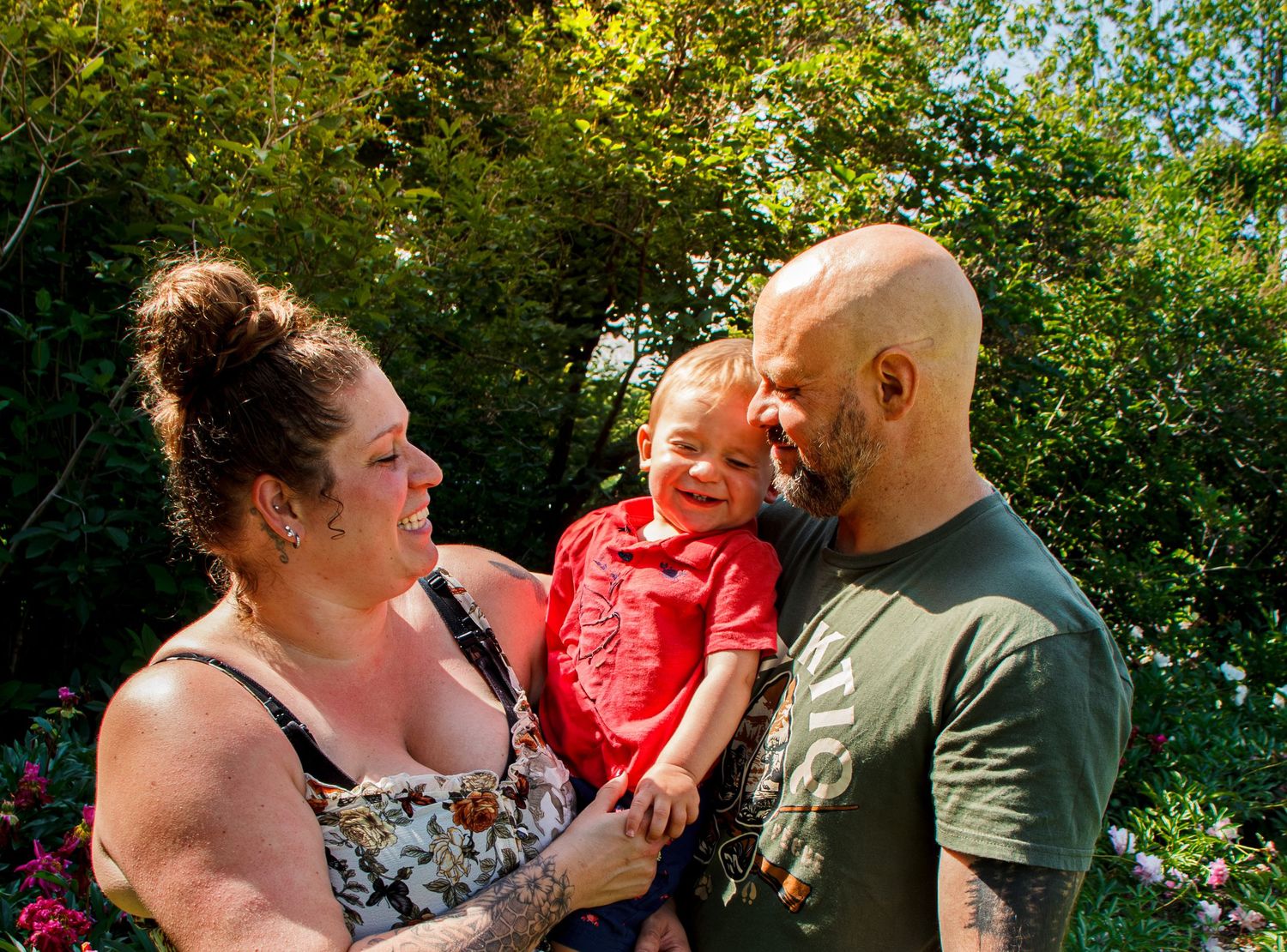 A smiling couple holds their laughing toddler in a red shirt outdoors surrounded by lush greenery.