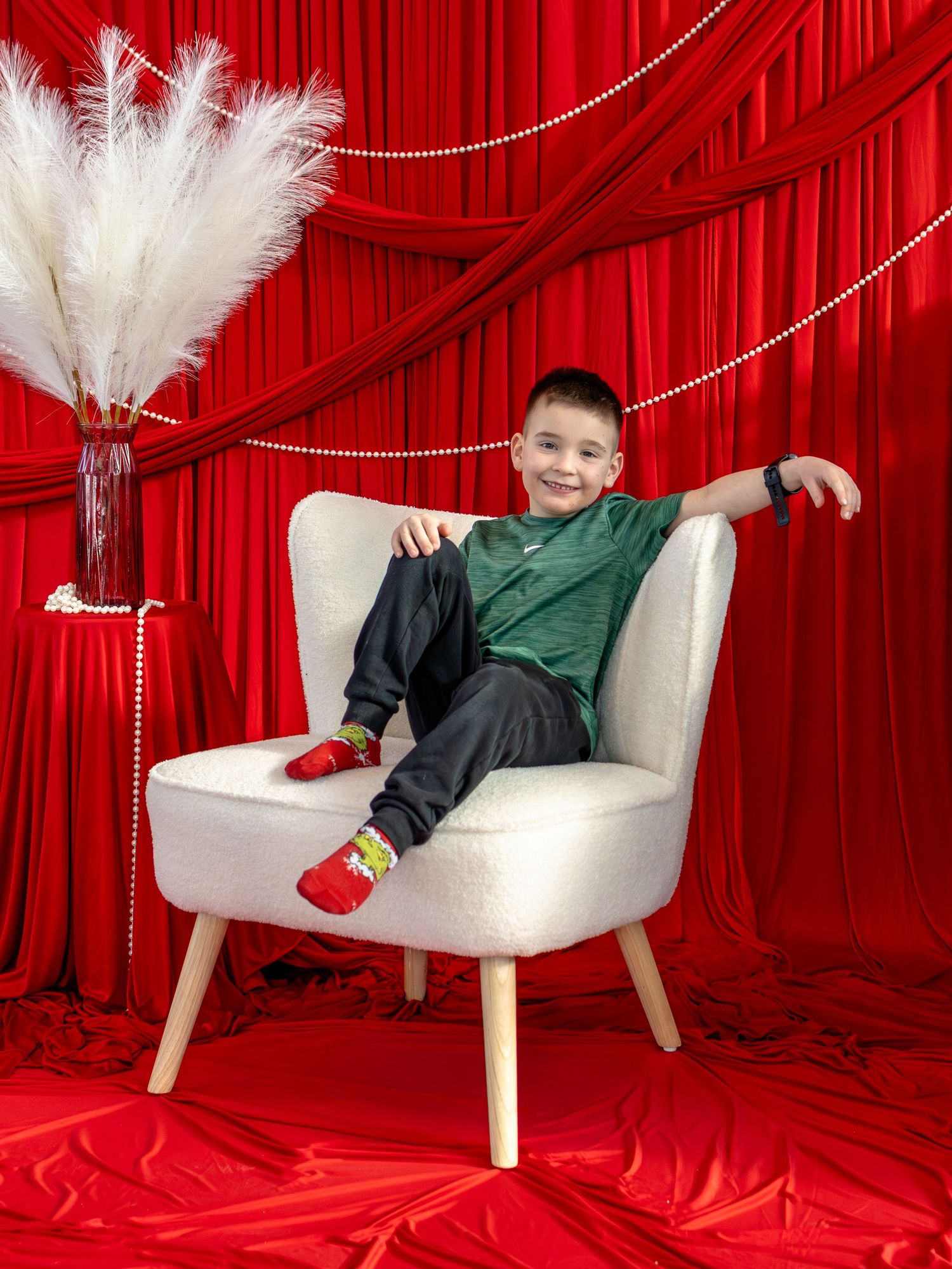 Smiling boy in green shirt sits relaxed in white chair against red velvet curtain backdrop with white feather decor.