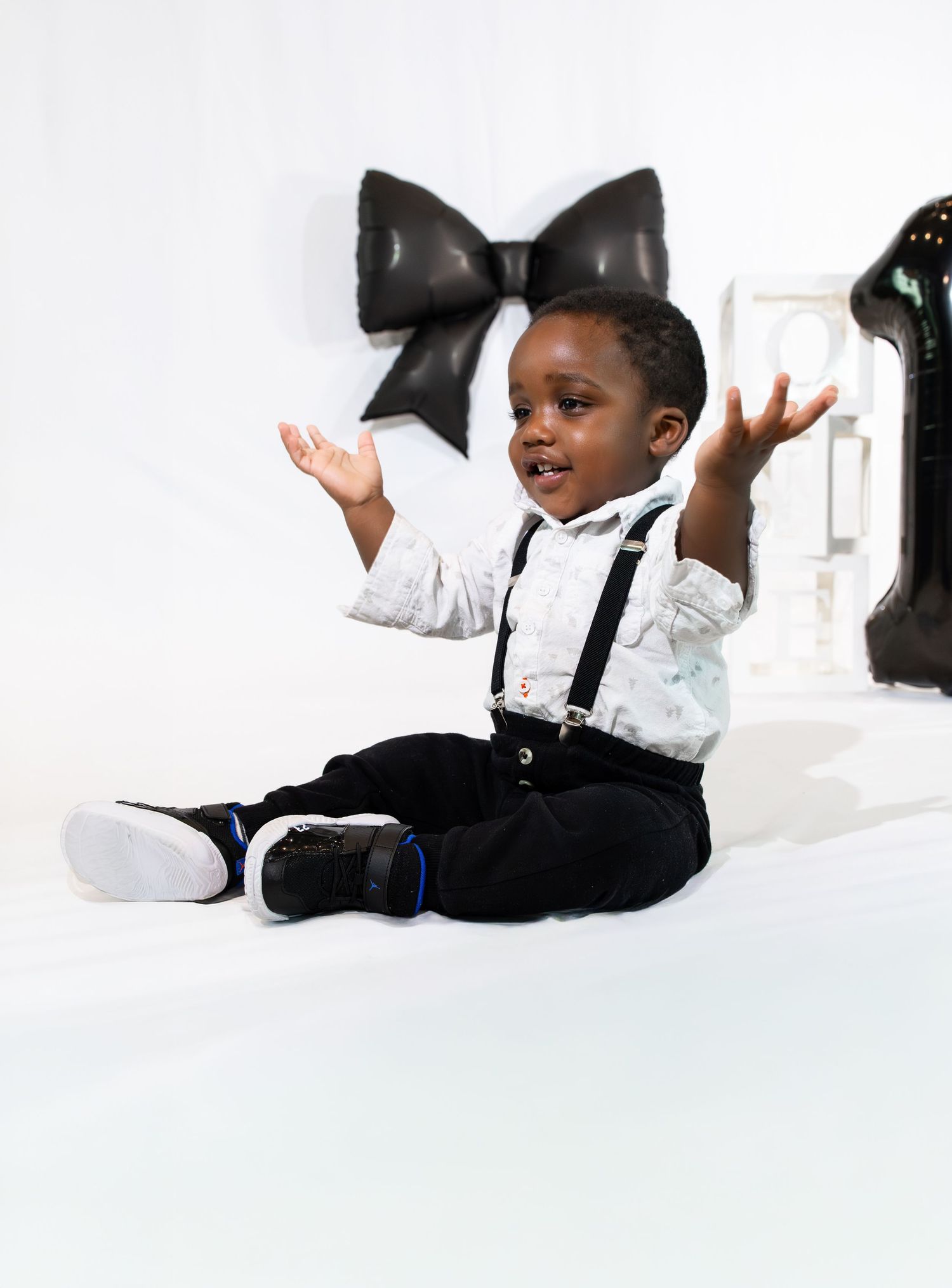 A joyful toddler in suspenders and dress shirt sits on white floor, hands raised, beside a large black bow and number 1.