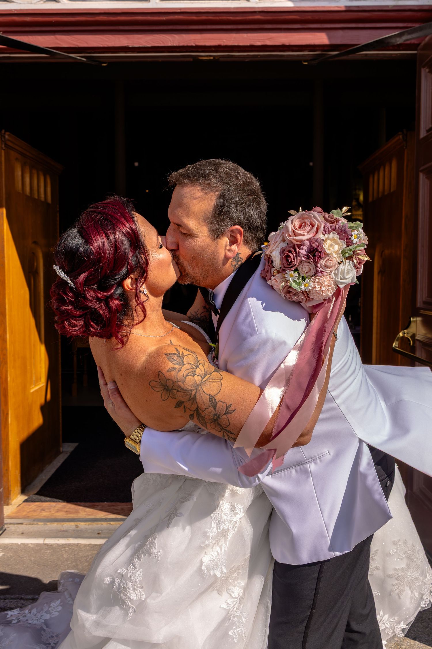 Bride and groom share a romantic kiss at church entrance, bride holding pink rose bouquet with visible arm tattoo.