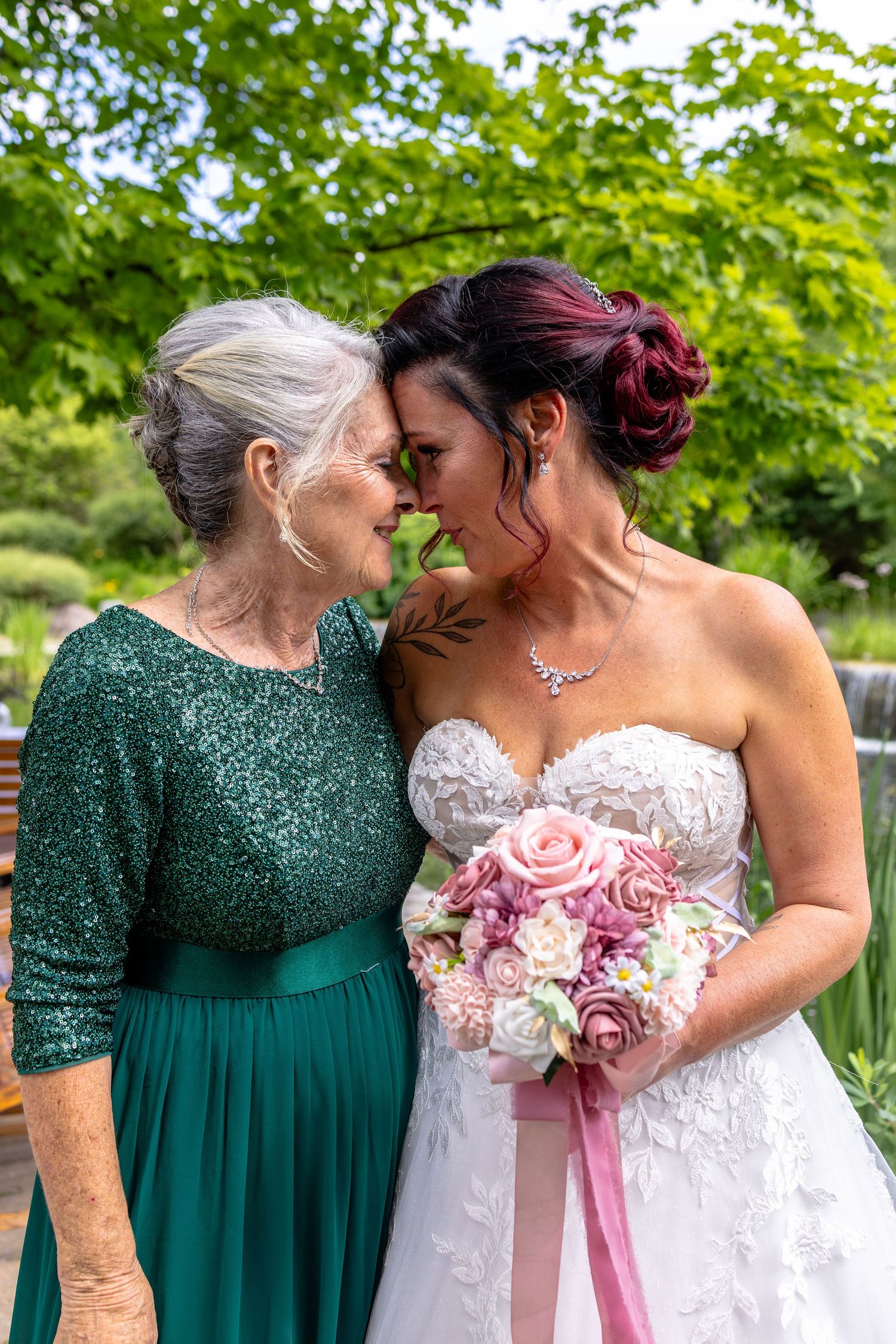 Bride in white gown holds pink bouquet, touching foreheads with elderly woman in green sequin dress outdoors.