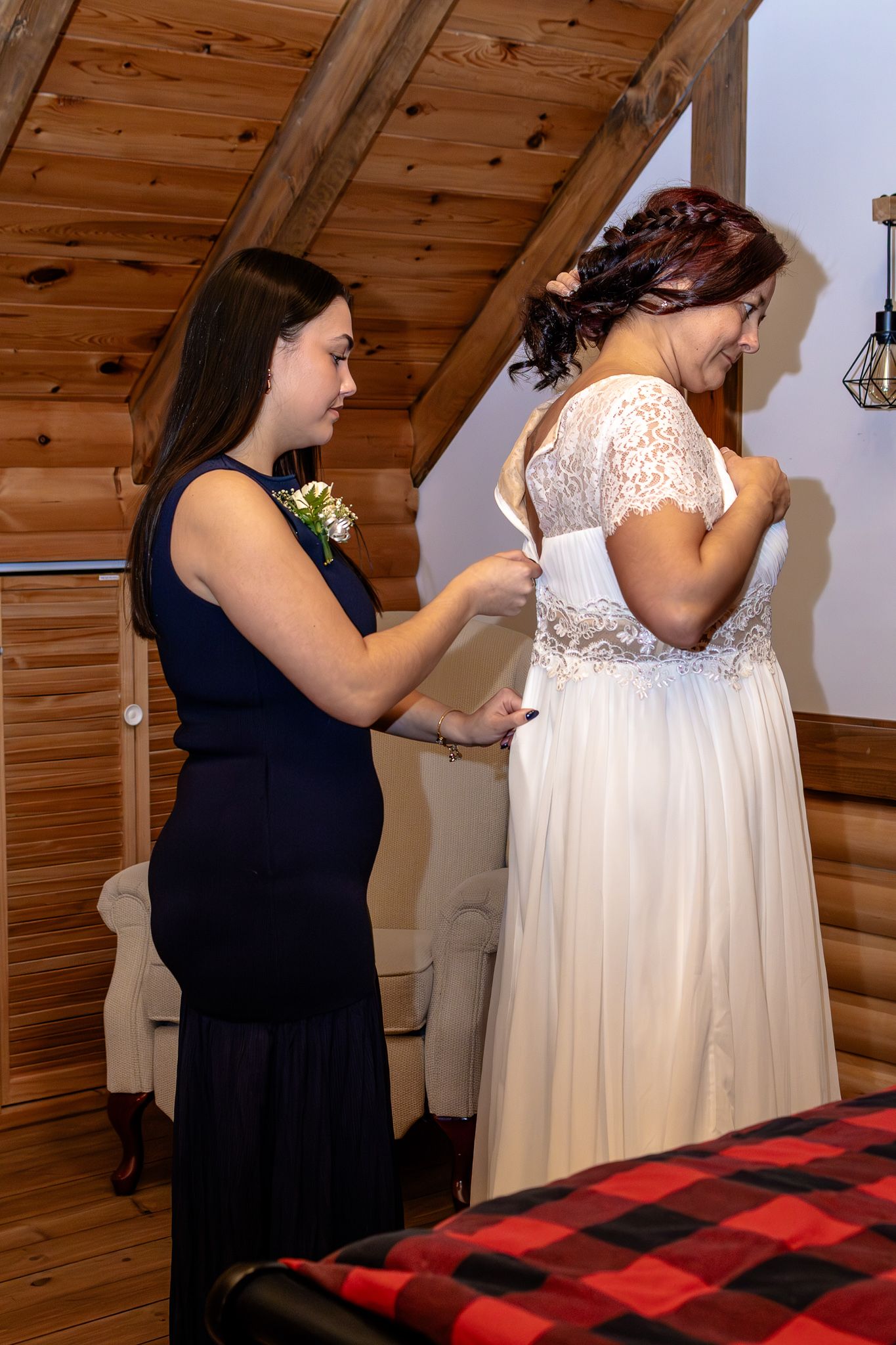 Bridesmaid in navy dress helping to fasten the back of a bride's white lace wedding gown in a log cabin.
