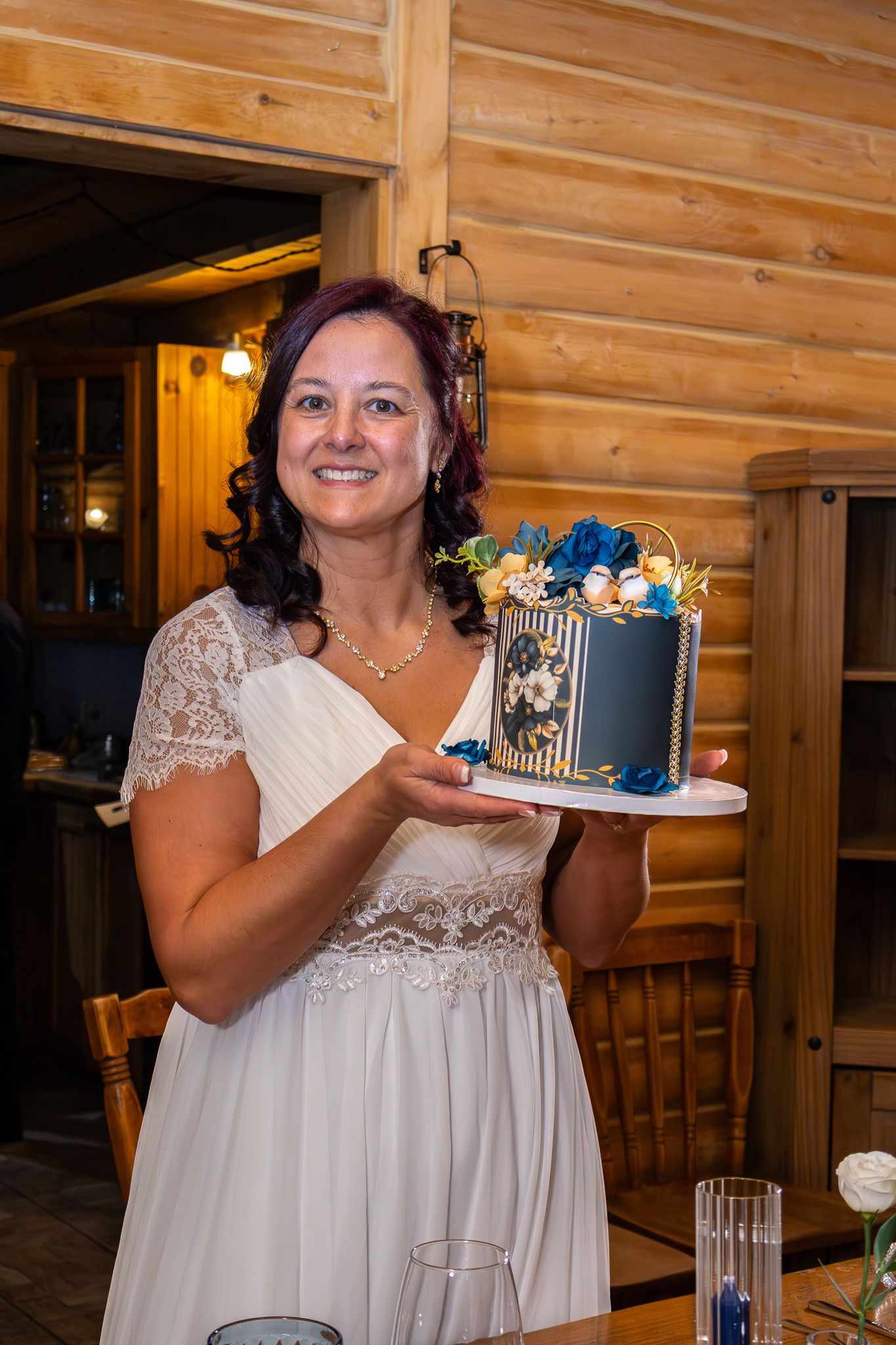 Smiling bride in white lace dress proudly displays an elegant navy and gold floral wedding cake.