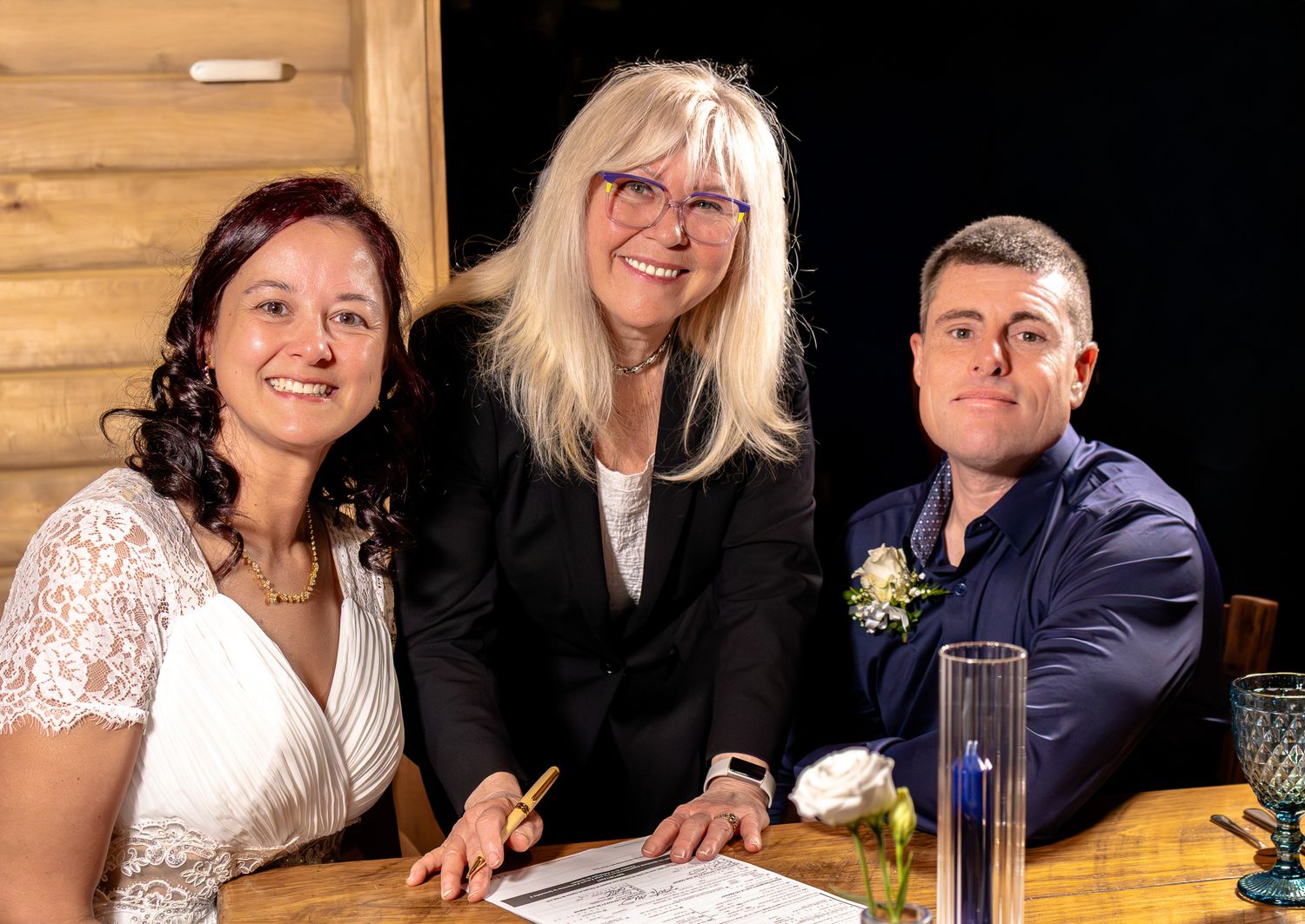 Bride, officiant, and groom smile together while signing wedding certificate at rustic wooden venue.