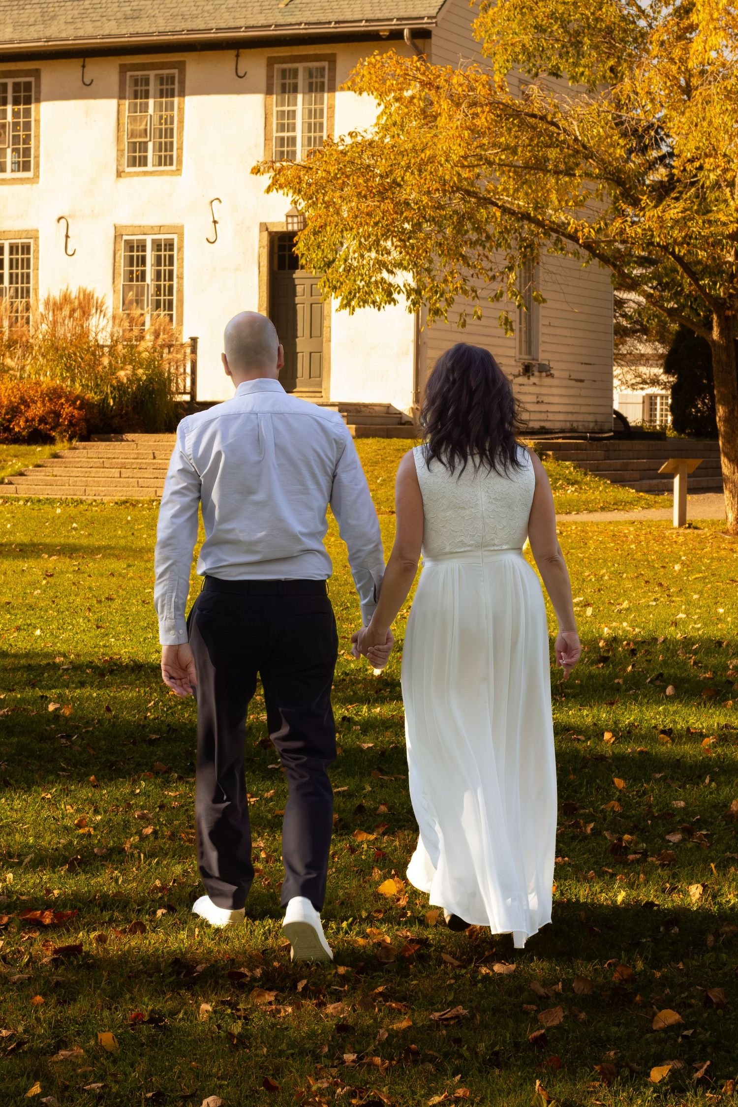 Bride and groom walking hand in hand toward a white historic building surrounded by golden autumn foliage.