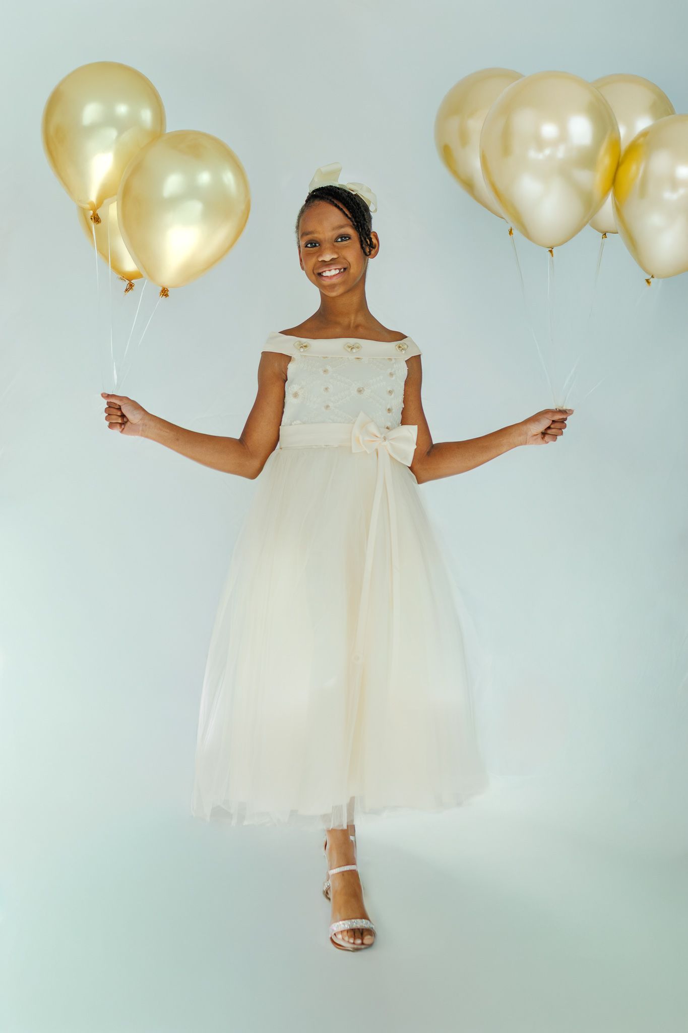 Smiling young girl in white tulle communion dress holding gold balloons on a light blue background.