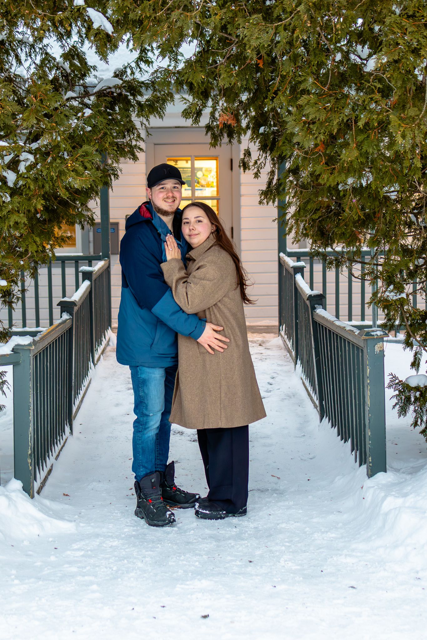 Couple embracing on snowy winter bridge surrounded by evergreen trees near a warmly lit home.