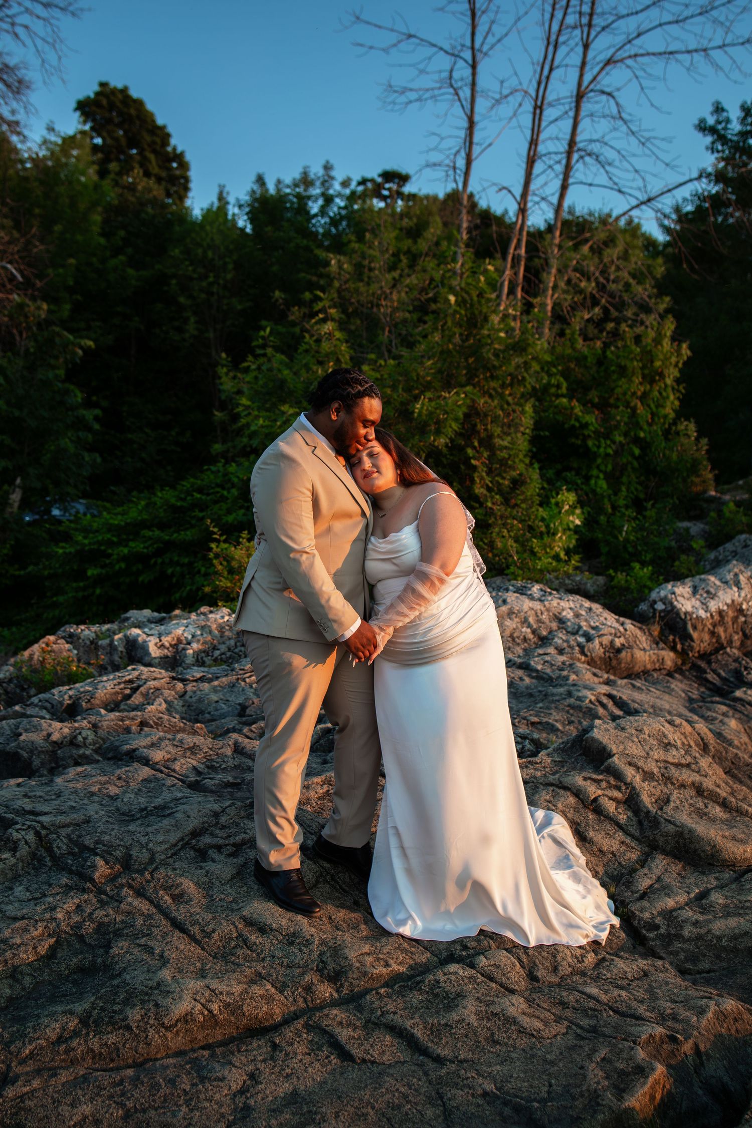 Newlywed couple embracing on rocky outcrop at golden hour, bride in white gown and groom in beige suit, forest backdrop.