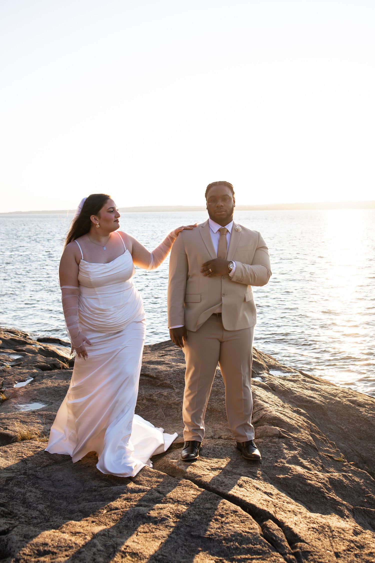 Bride and groom pose on rocky shoreline at sunset, she in white gown, he in tan suit, water behind them.