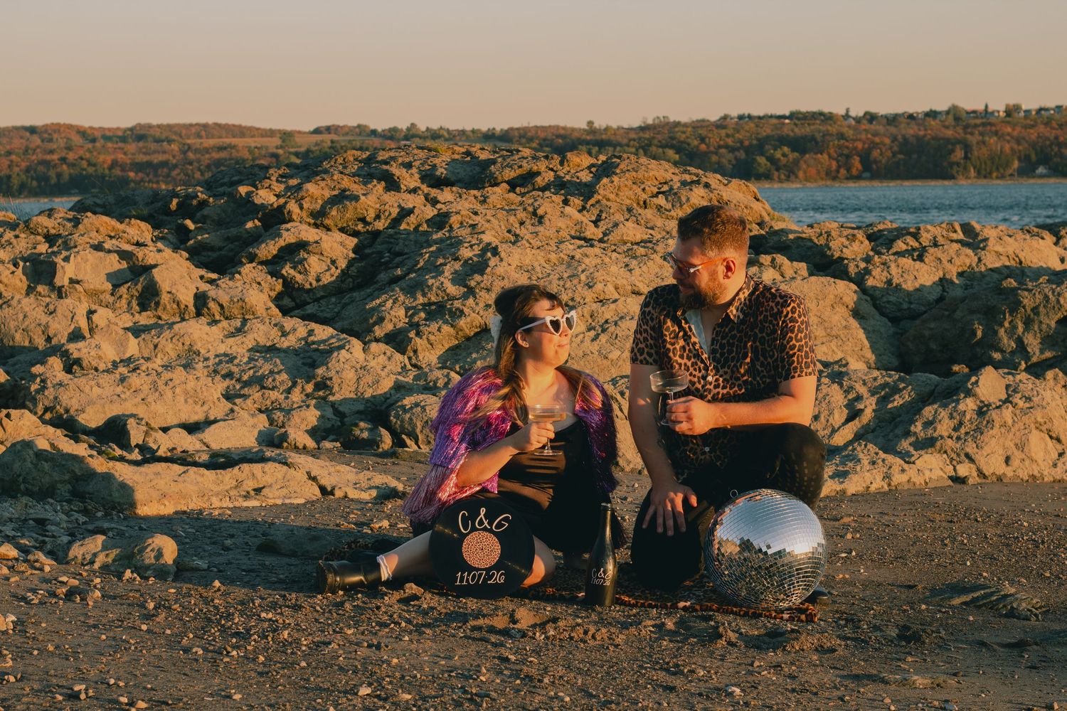 Two people sitting on rocky shore at golden hour with a mirrored ball beside them.