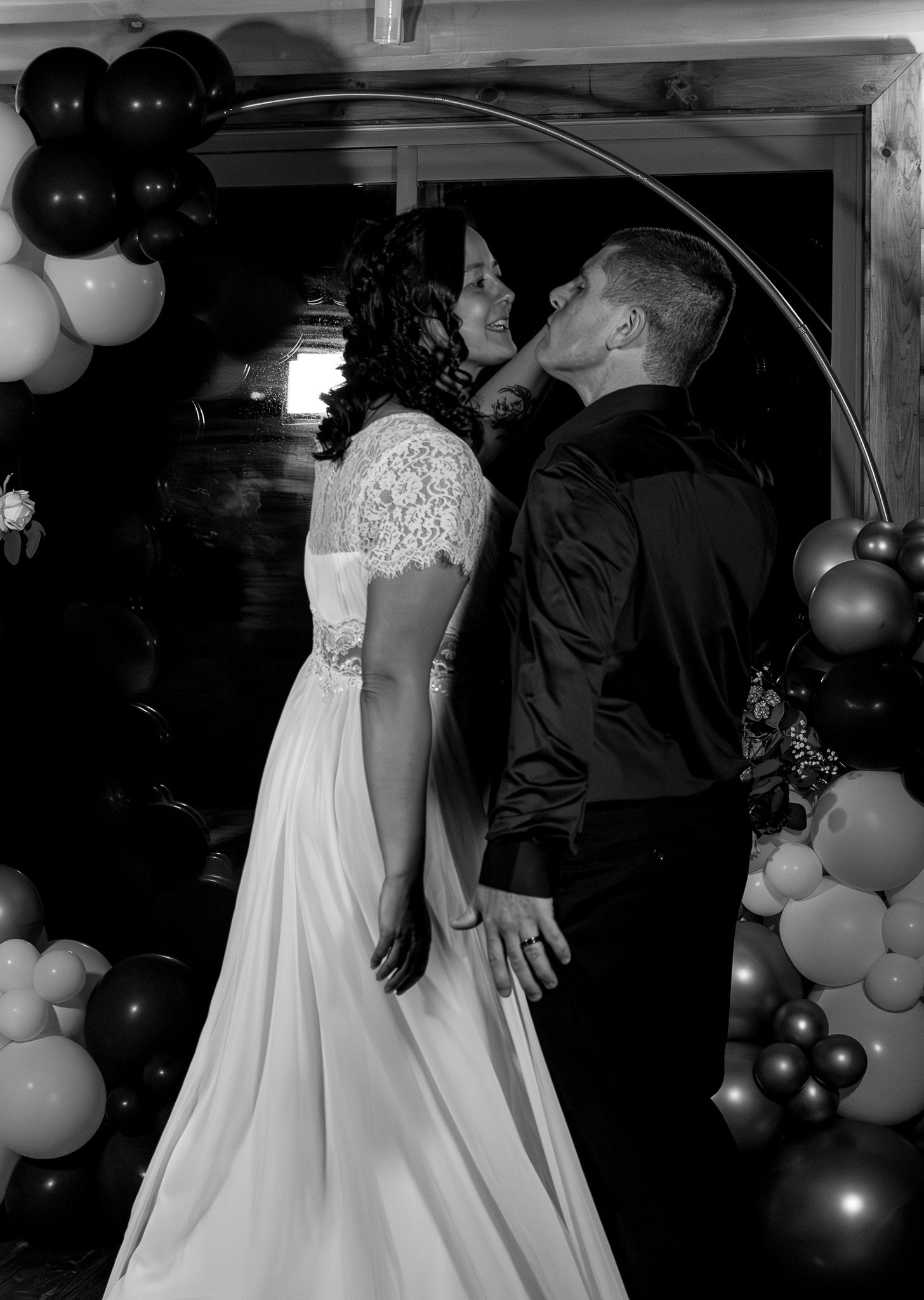 Black and white photo of bride and groom sharing a kiss under circular arch with balloon decorations.