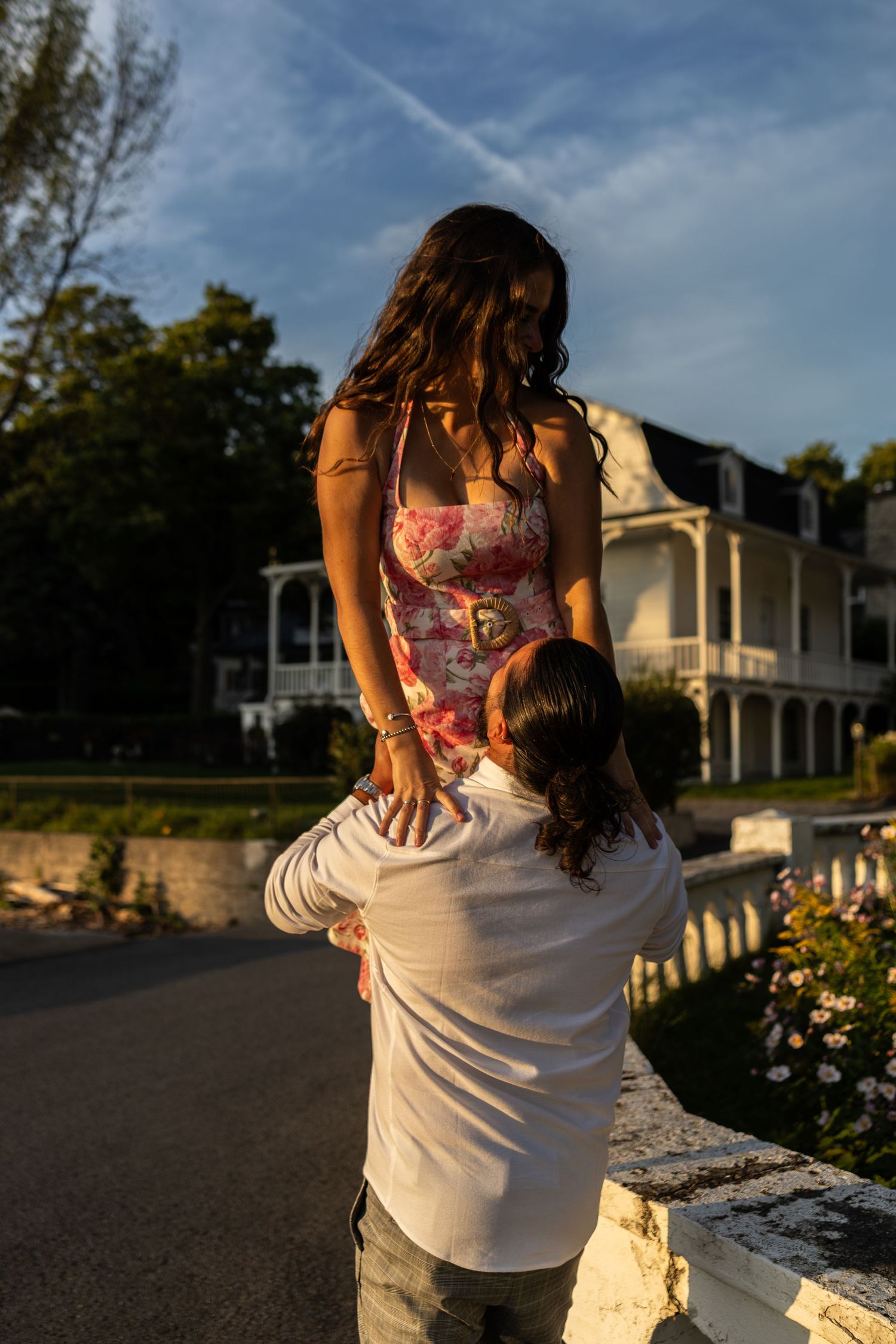 A man lifts a woman in a floral dress outdoors near a historic house at golden hour.