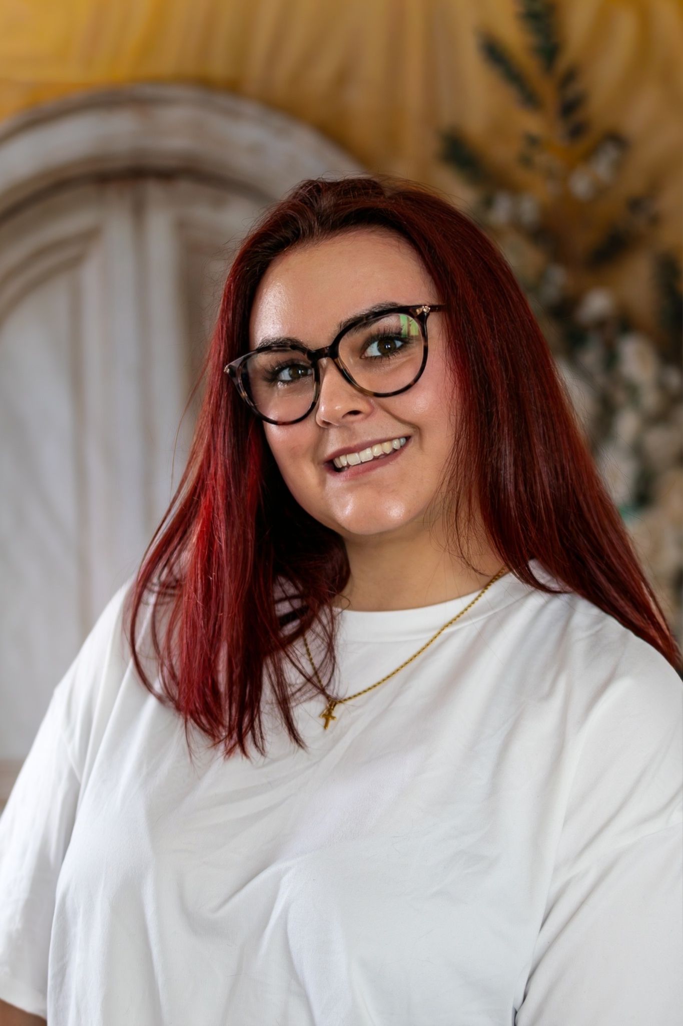 Smiling young woman with red hair and glasses wears white tee against decorative backdrop.