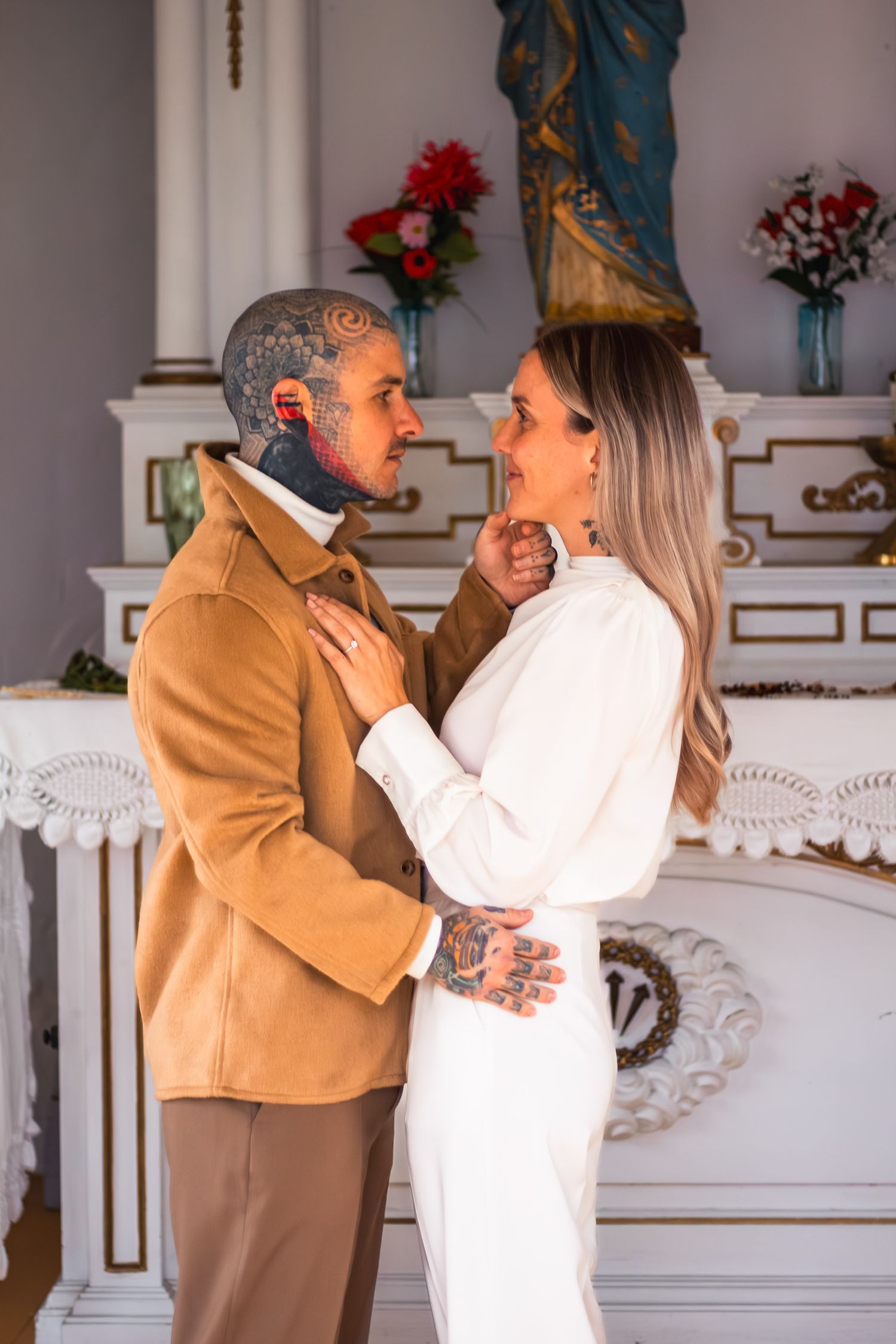 Tattooed groom and smiling bride embrace tenderly in front of an ornate white altar with flowers.