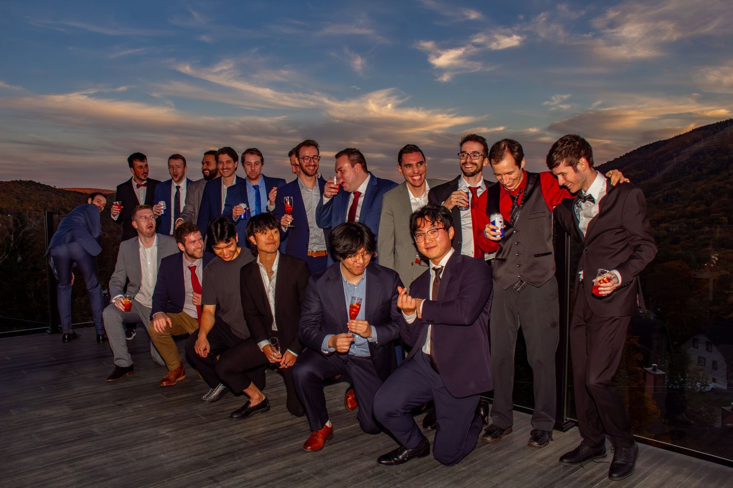 Large group of male wedding guests pose and celebrate together outdoors during a vibrant purple sunset.