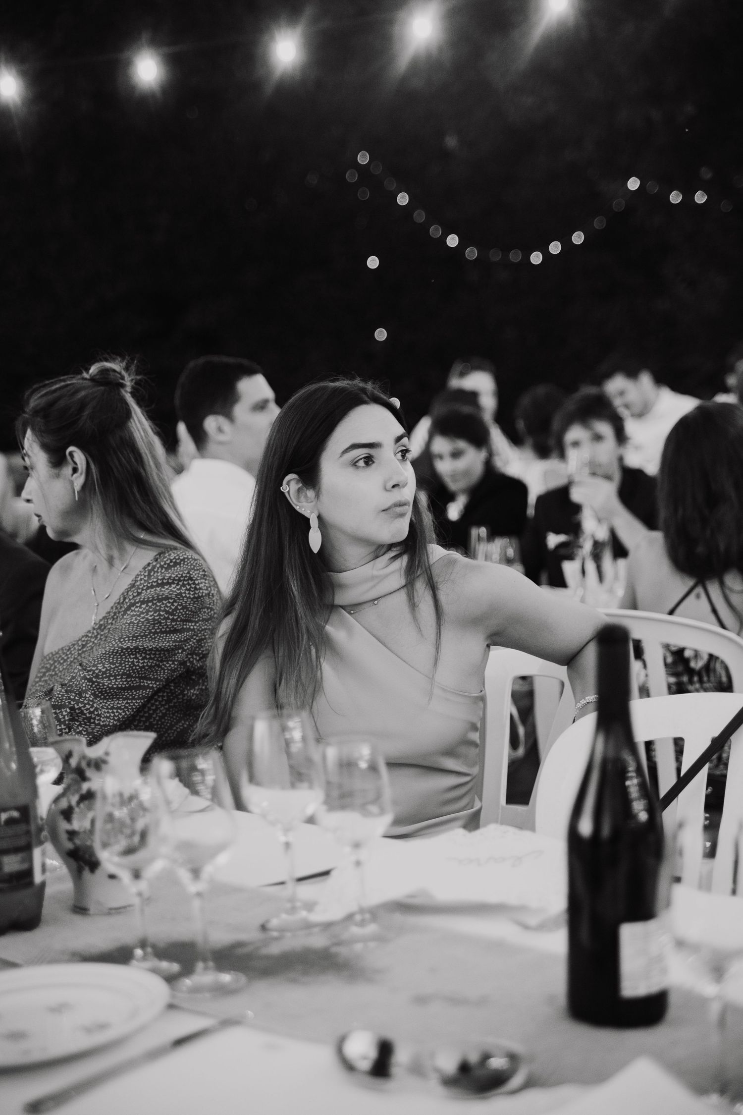 Black and white photo of diners at an elegant dinner party with wine glasses and string lights overhead.