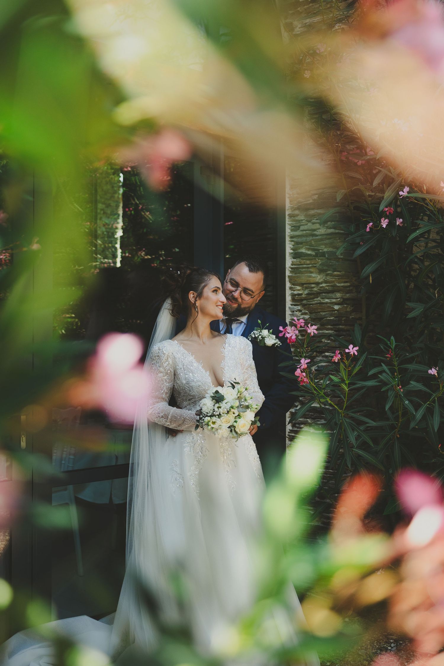 Romantic garden wedding photo with couple embracing among pink flowers and greenery through soft focus lens.