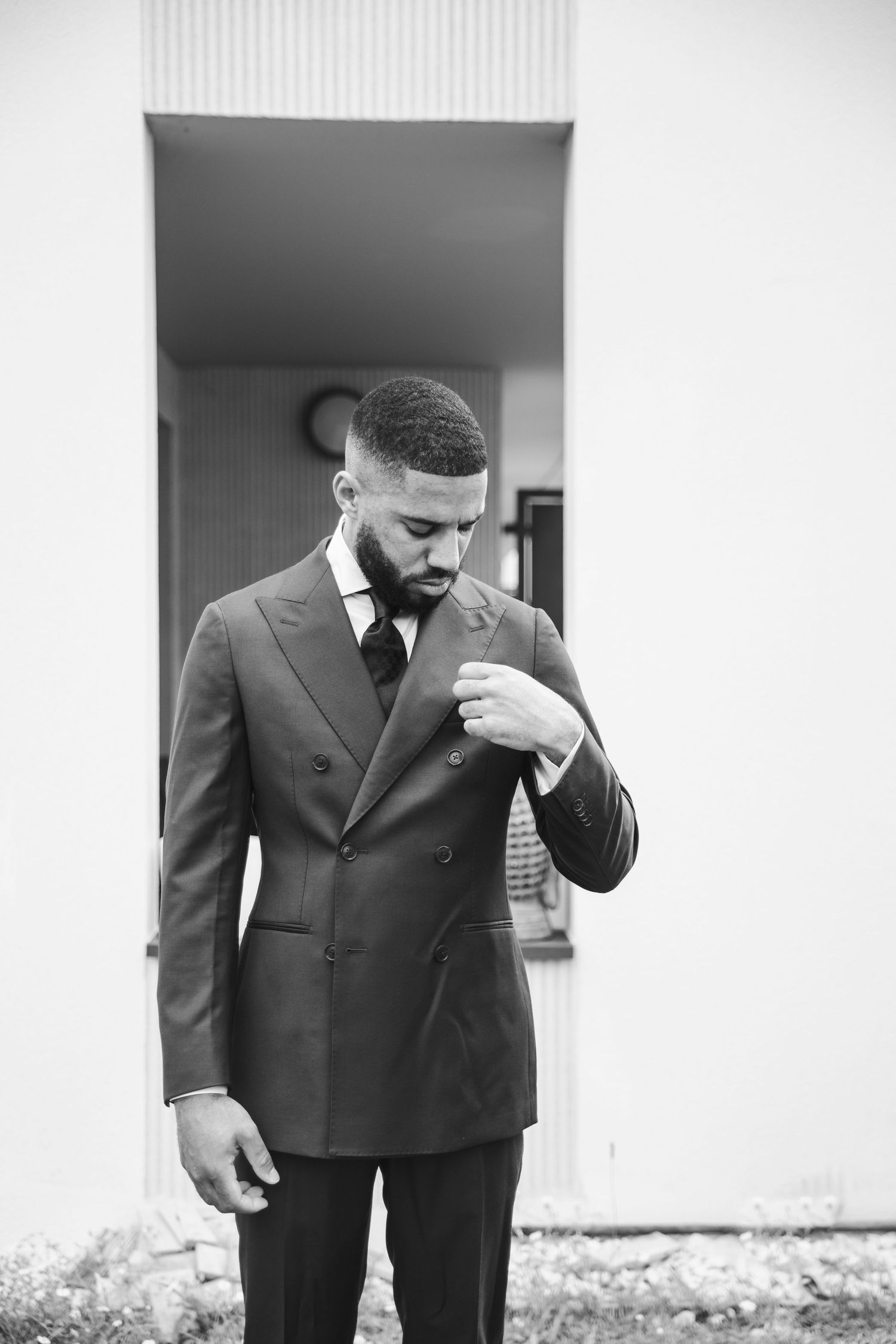 A well-dressed person in a double-breasted suit looks down while adjusting their collar in a black and white portrait.