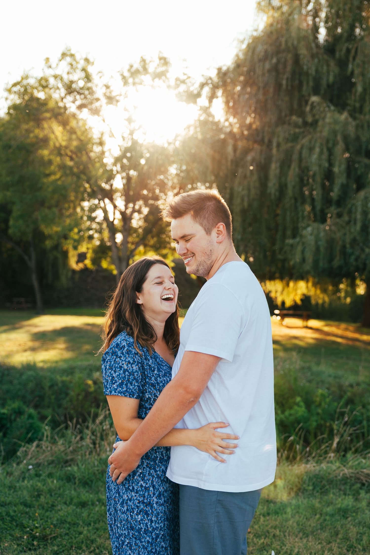 A couple shares a joyful moment while embracing outdoors at sunset with weeping willow trees in the background.