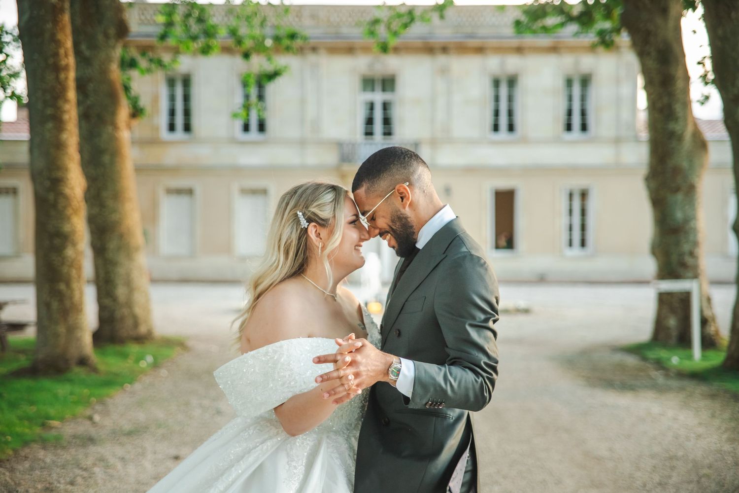 A couple in wedding attire share a romantic moment in front of a French chateau on a tree-lined path.